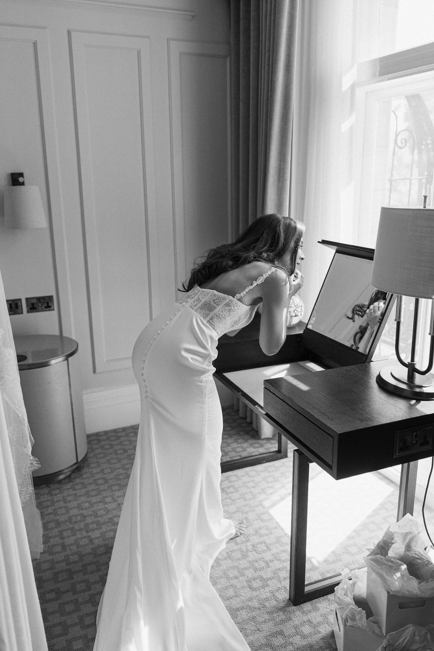 Black and white photo of bride in fitted wedding gown leaning over dressing table by window.