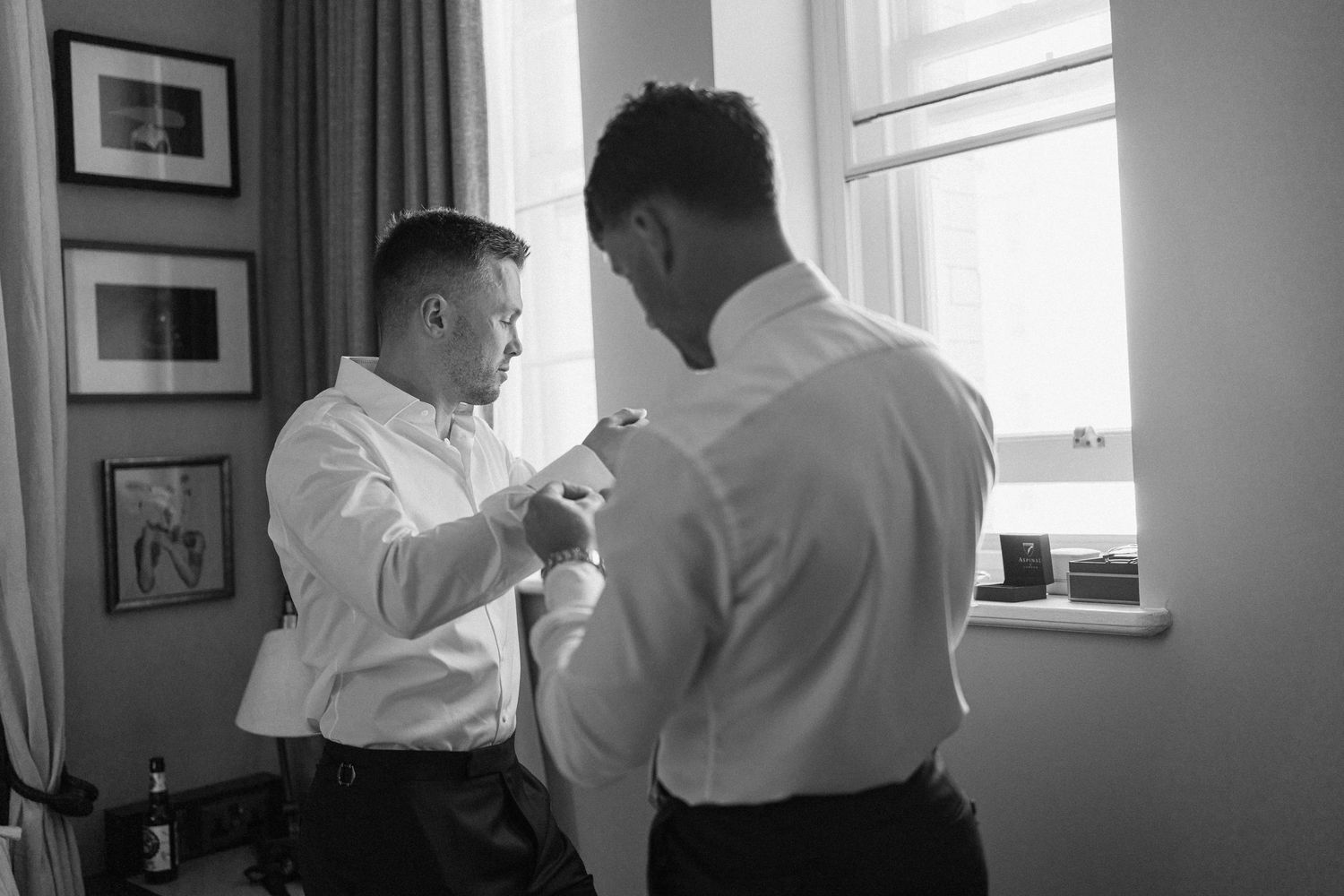 Two groomsmen in white dress shirts helping each other prepare before a wedding ceremony in a classic room.