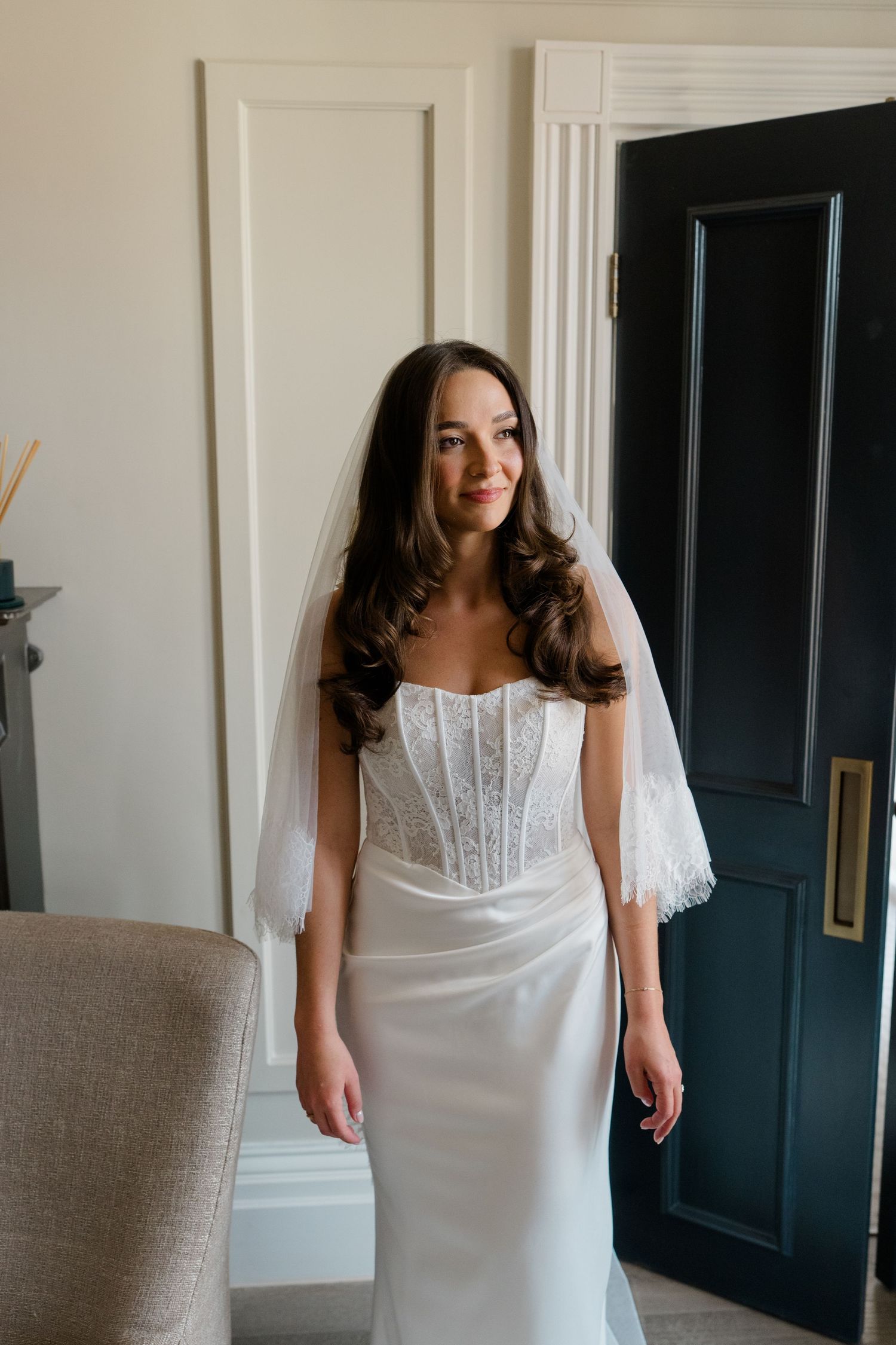 Smiling bride in white corset lace wedding gown and veil standing by a dark elegant door.
