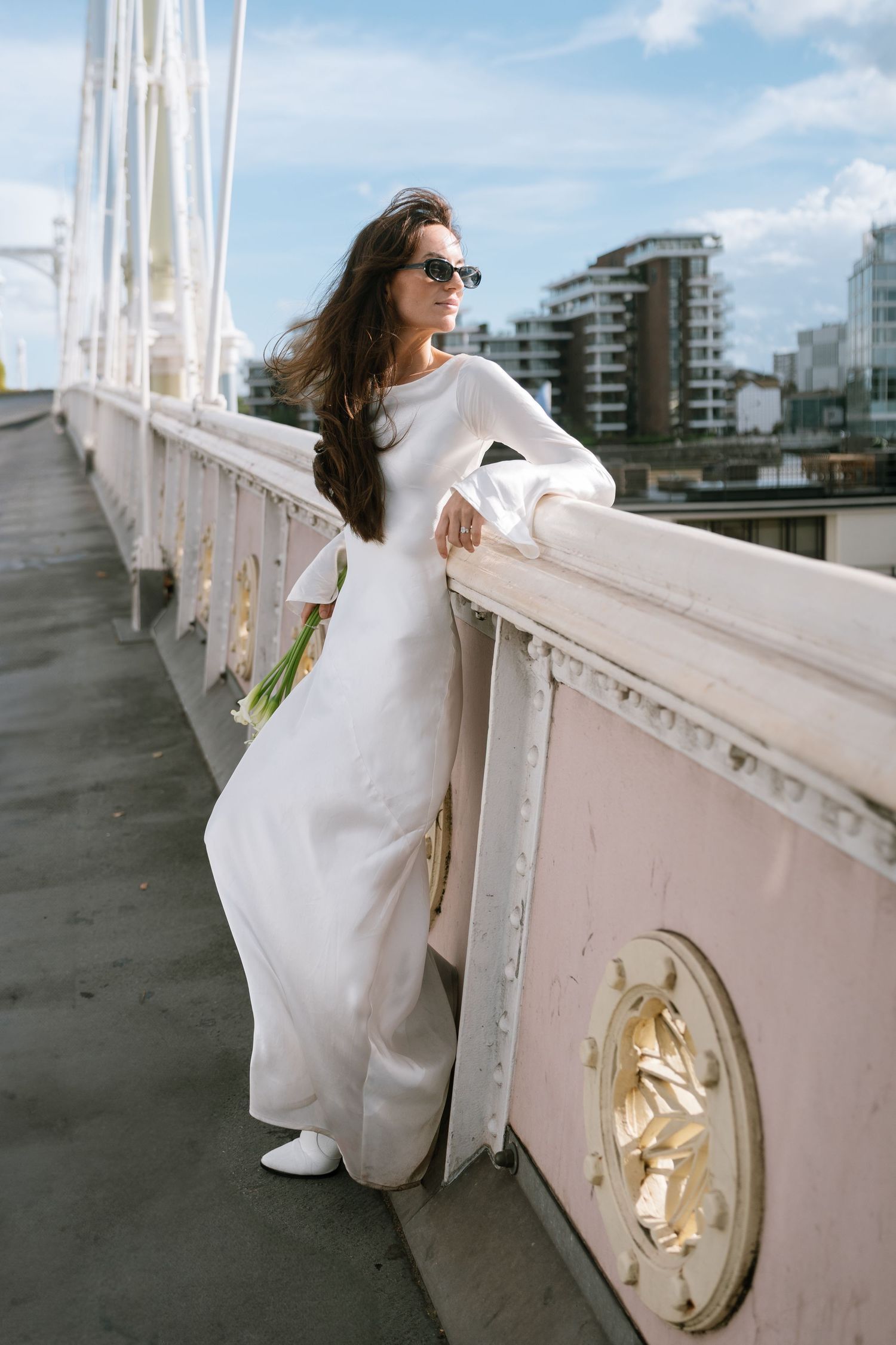 Woman in flowing white dress poses along a railing with city buildings in background.
