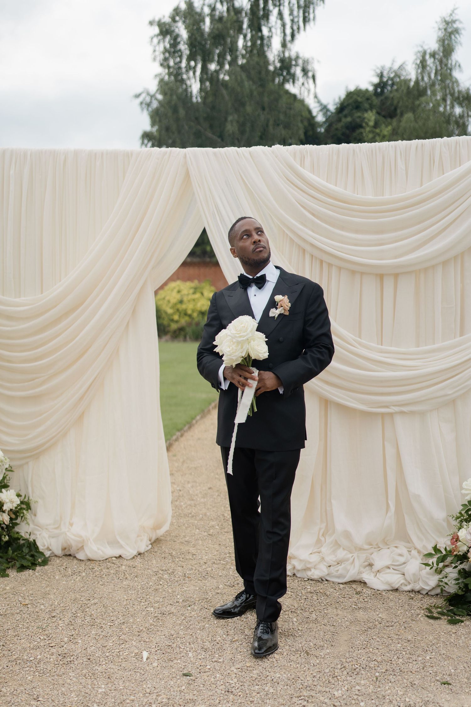 Handsome Black groom in tuxedo holding white bouquet standing at elegant draped wedding ceremony arch.