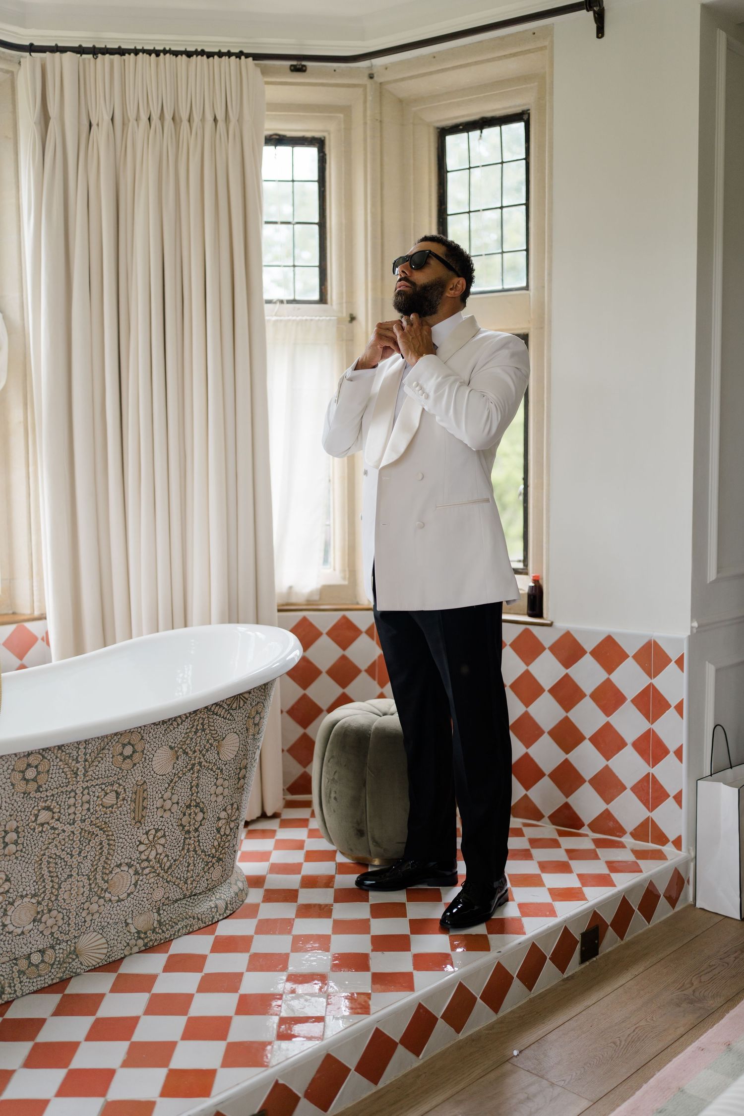 Groom in white tuxedo jacket adjusting bow tie in bathroom with red checkered tile floor.
