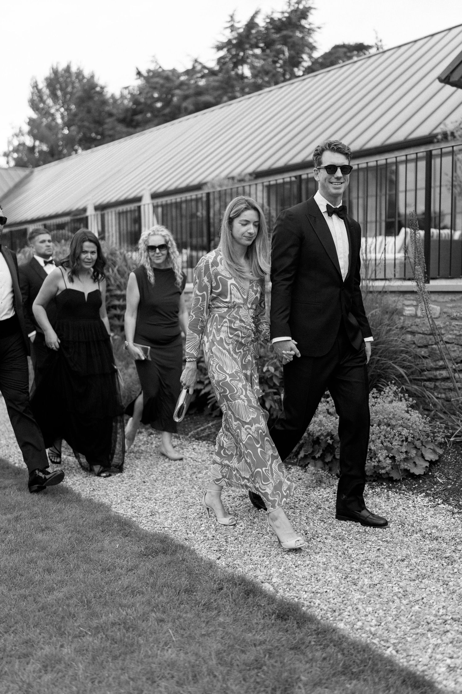 Elegantly dressed wedding guests walk along a gravel path in black and white photography.