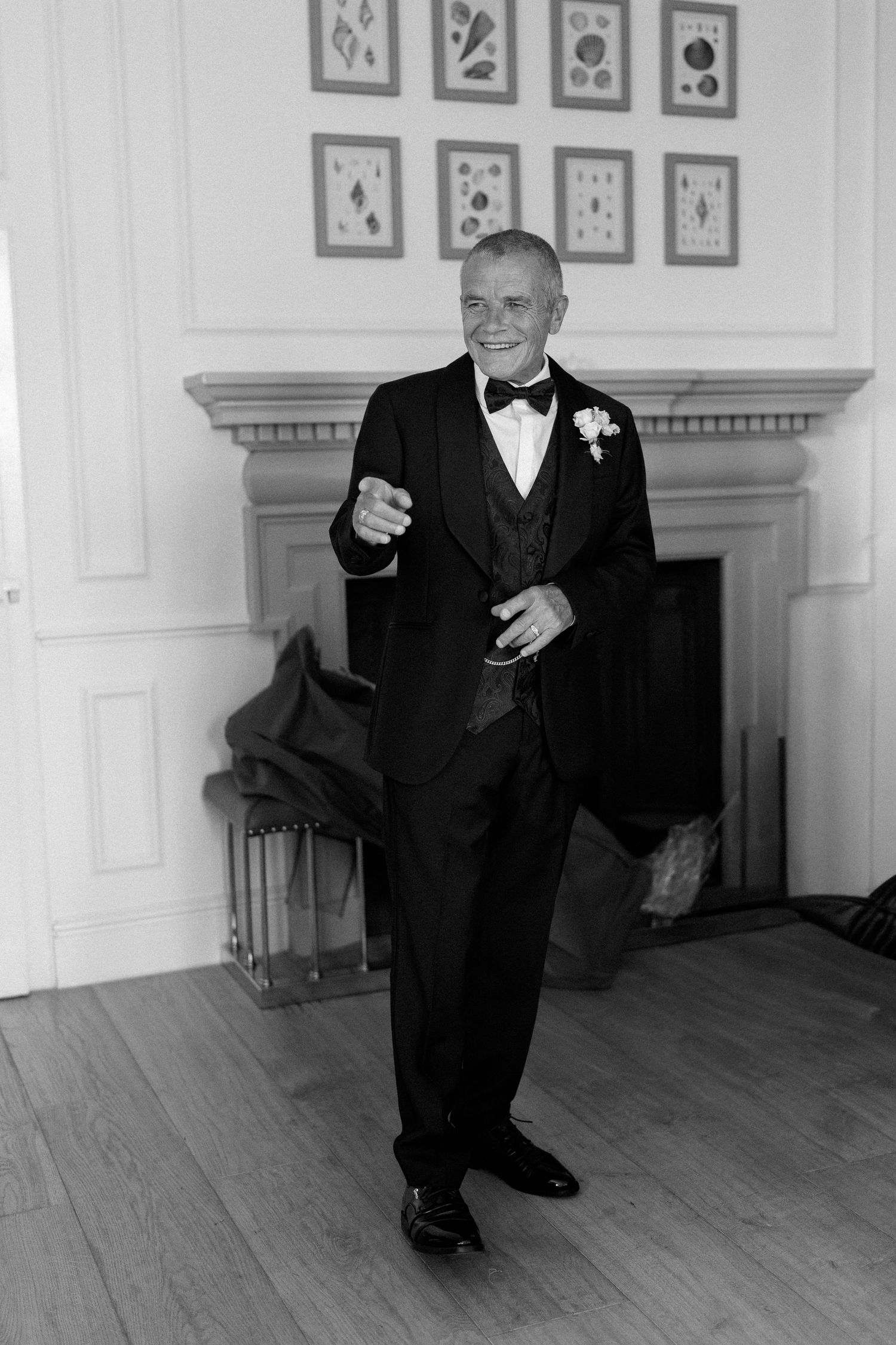 Dapper older man in black tuxedo with boutonniere posing confidently by a fireplace in black and white.