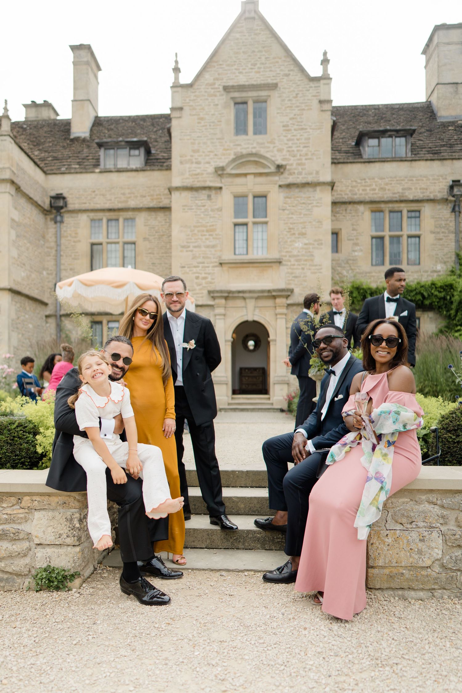 A group of wedding guests in formal wear pose on steps in front of a grand stone manor.