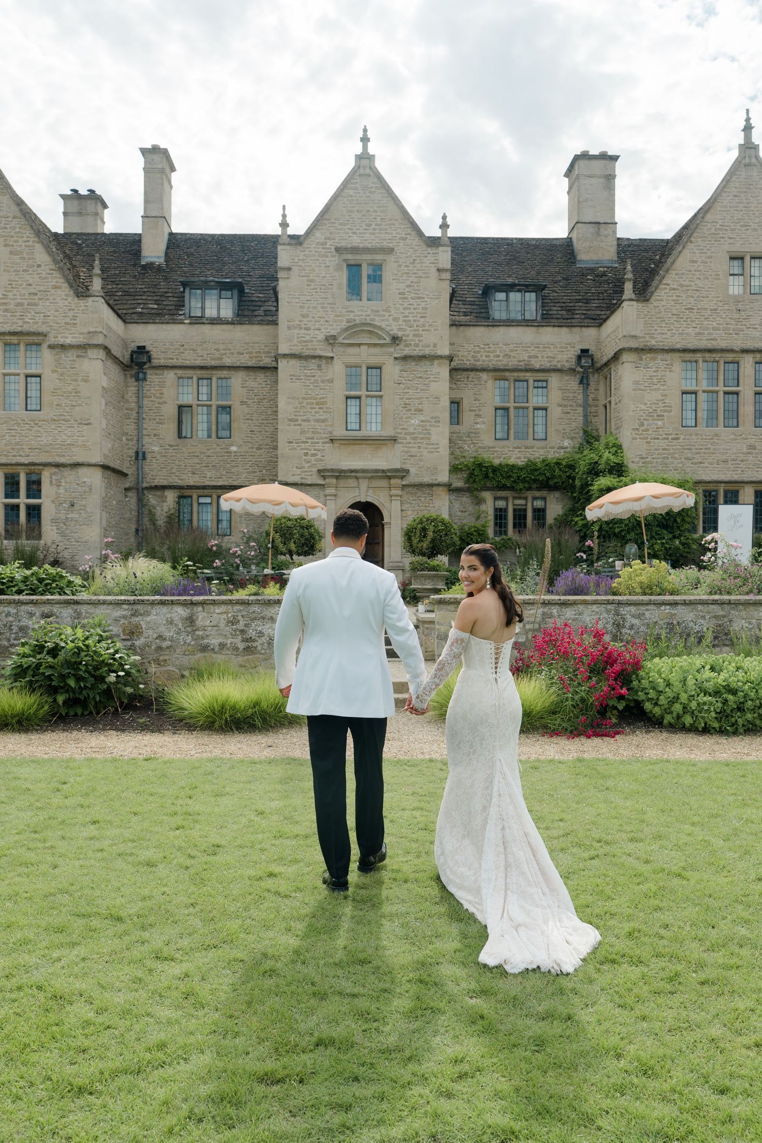 Bride and groom holding hands walking toward an elegant stone manor house with manicured gardens.