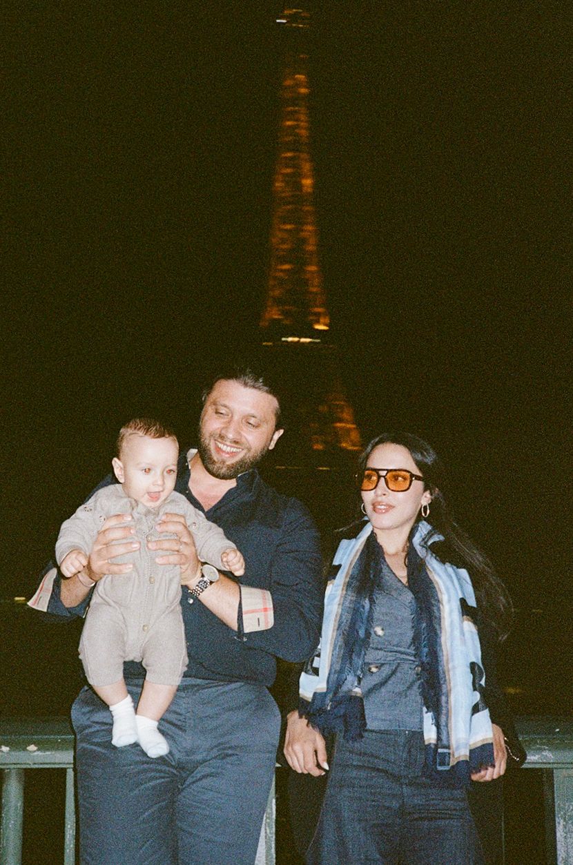 A smiling couple holding a baby poses in front of the illuminated Eiffel Tower at night in Paris.
