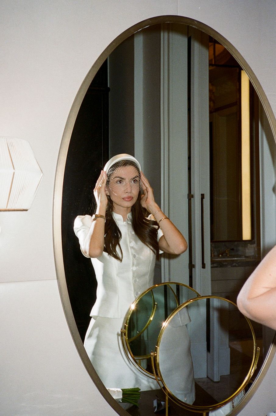Woman in white suit adjusting headband, reflected in large oval gold-framed mirror.