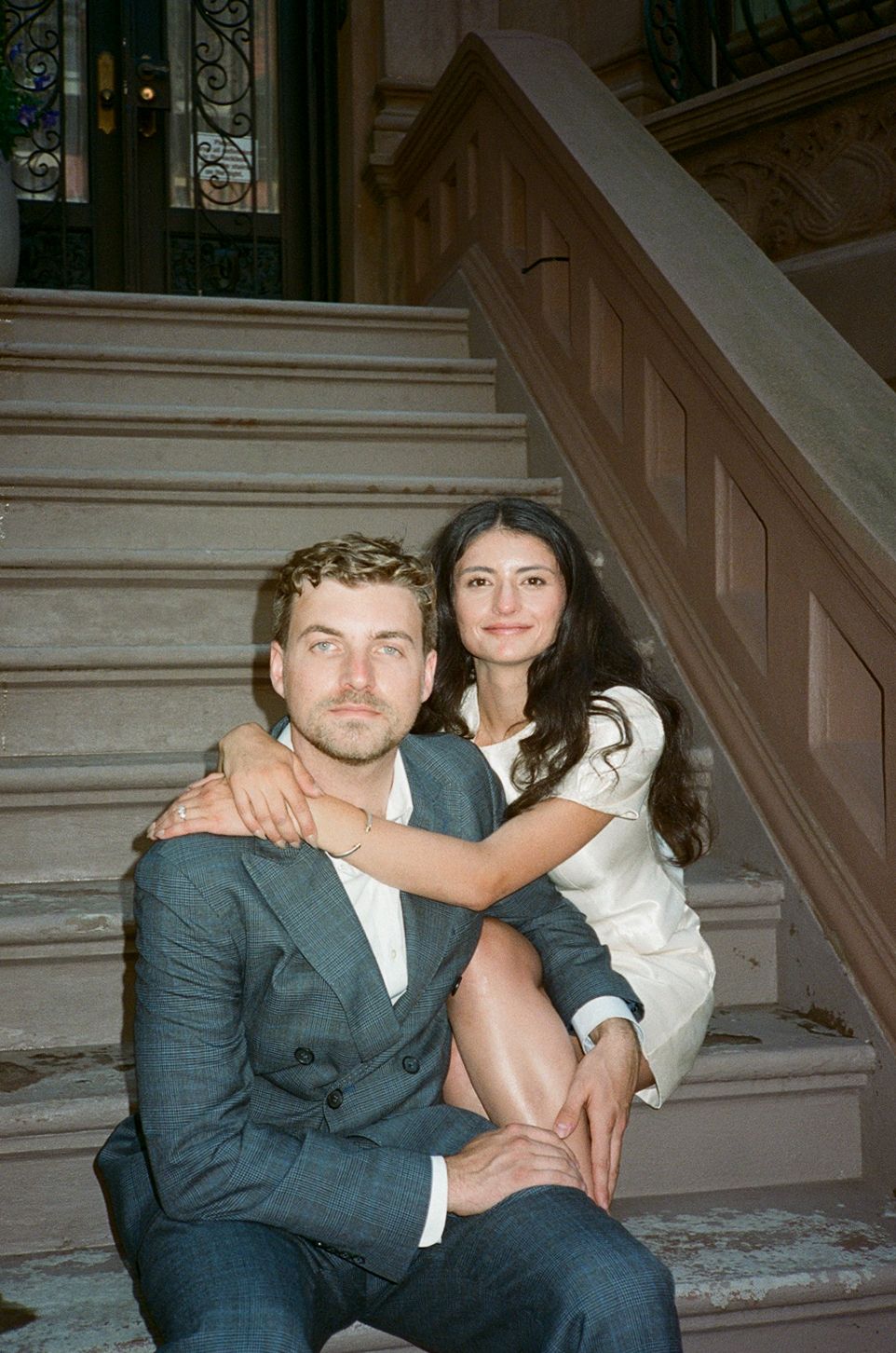 Couple posing on elegant staircase steps, man in gray suit seated, woman in white dress behind him with arm around his shoulder.