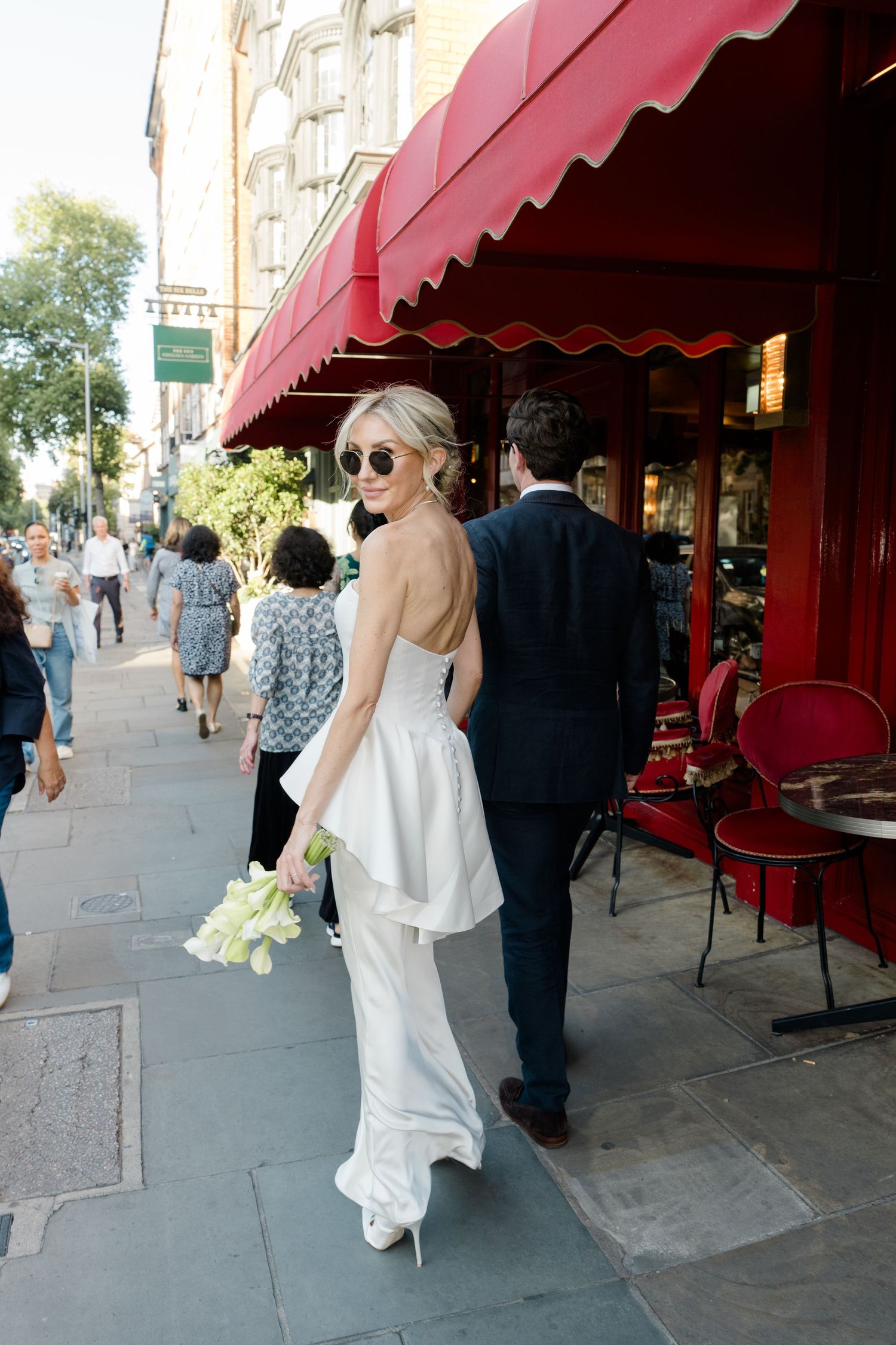 Bride in white strapless gown and sunglasses walks with groom past red awning restaurant on city sidewalk.