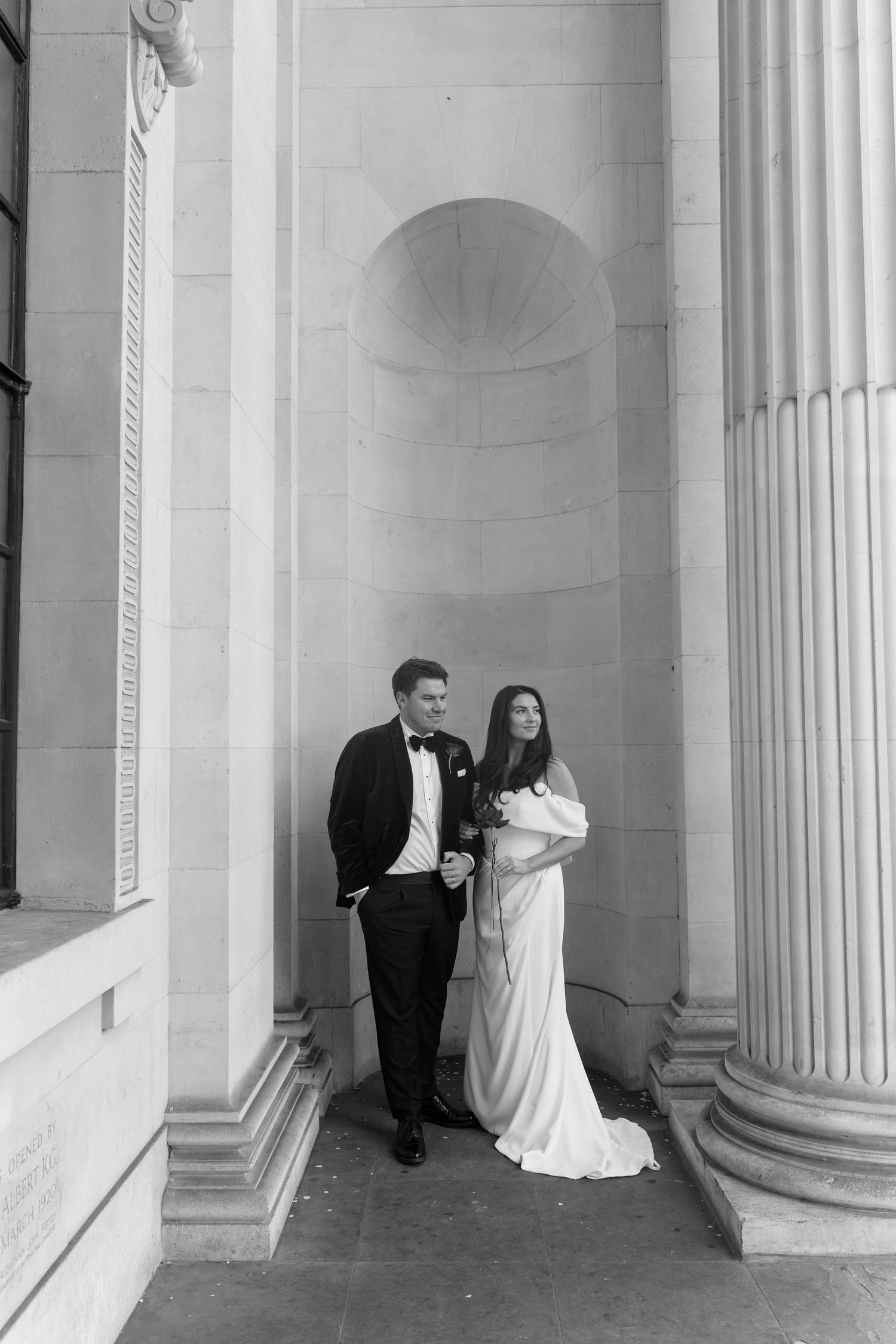 Black and white wedding portrait of a bride in an off-shoulder gown and groom in a tuxedo standing beside stone columns and an arched niche.