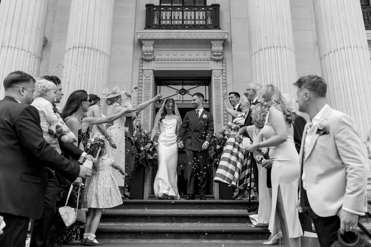 Black and white photo of a newlywed couple exiting a grand columned building as guests throw confetti.