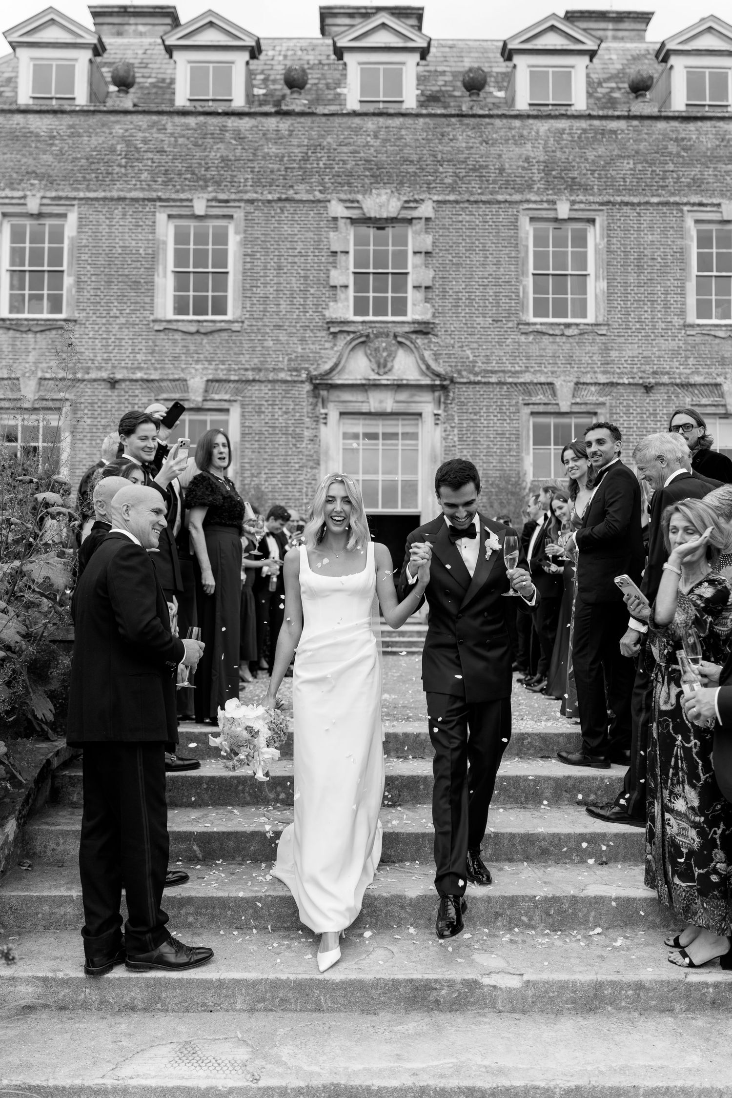 Black and white photo of bride and groom walking down steps amid confetti outside a grand brick manor house.