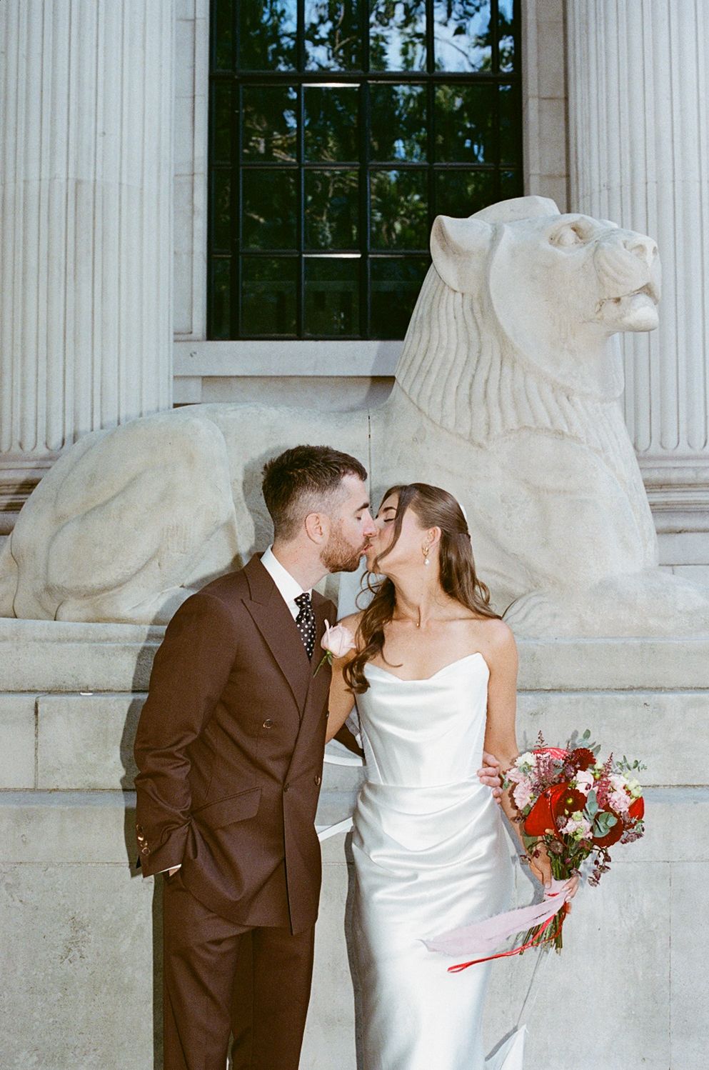 Bride and groom kissing in front of a stone lion sculpture and columns at a classic wedding venue.