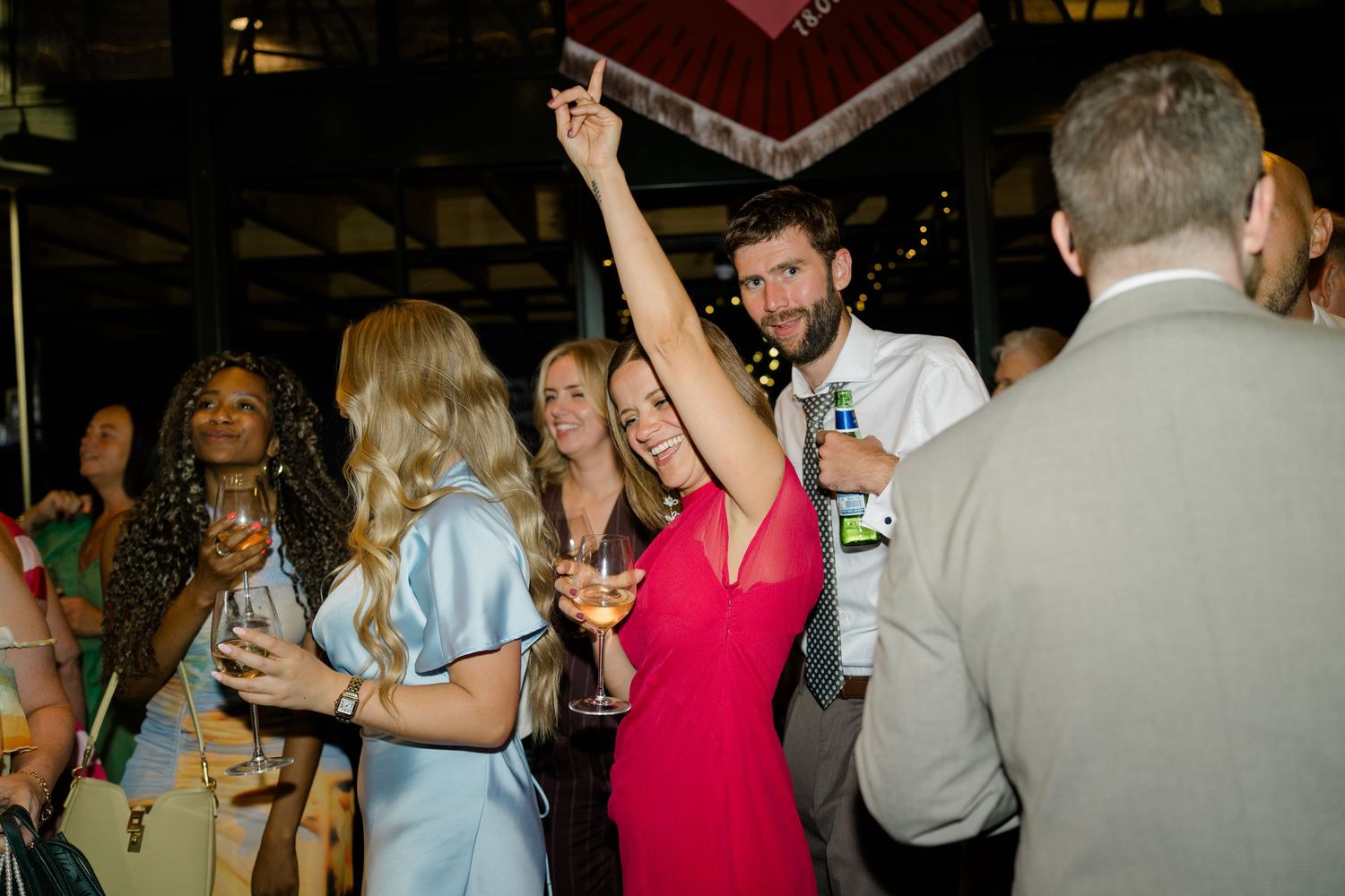 Energetic wedding guest in red dress raises hand on dance floor surrounded by celebrating guests at reception.