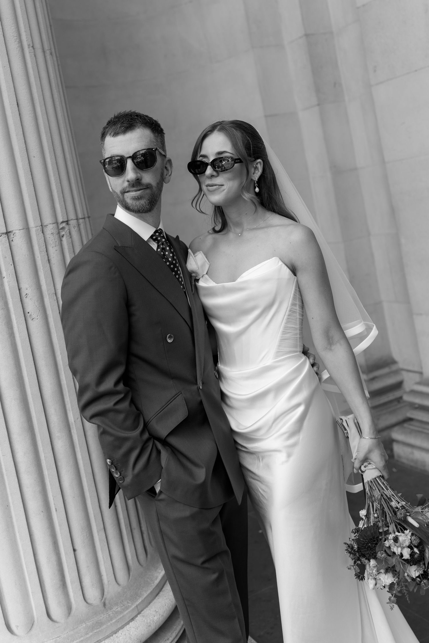 Stylish bride and groom in sunglasses posing by stone columns.