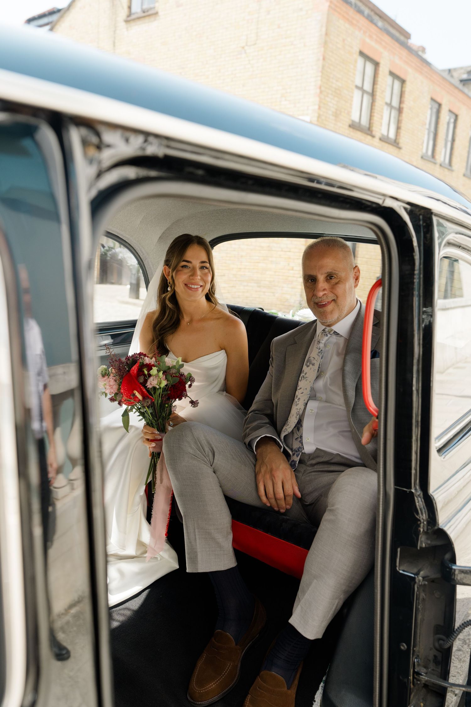 Bride and groom sitting in vintage black London taxi cab on wedding day with bridal bouquet.