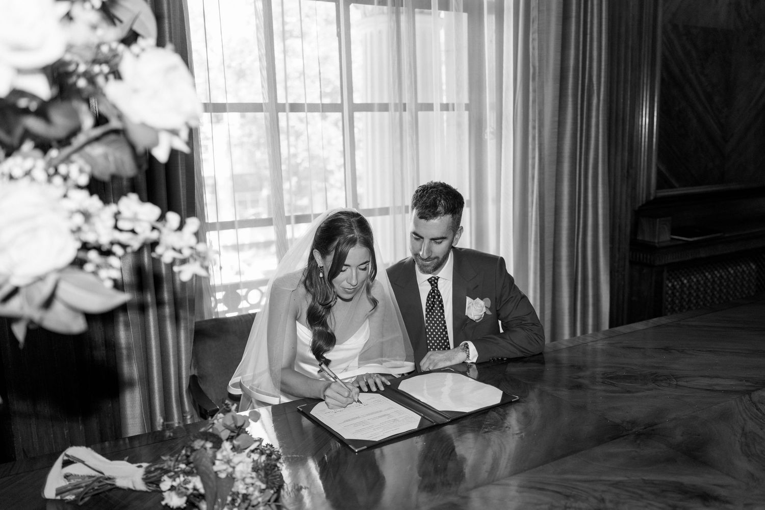 Bride and groom signing wedding register documents at a table in black and white photo.