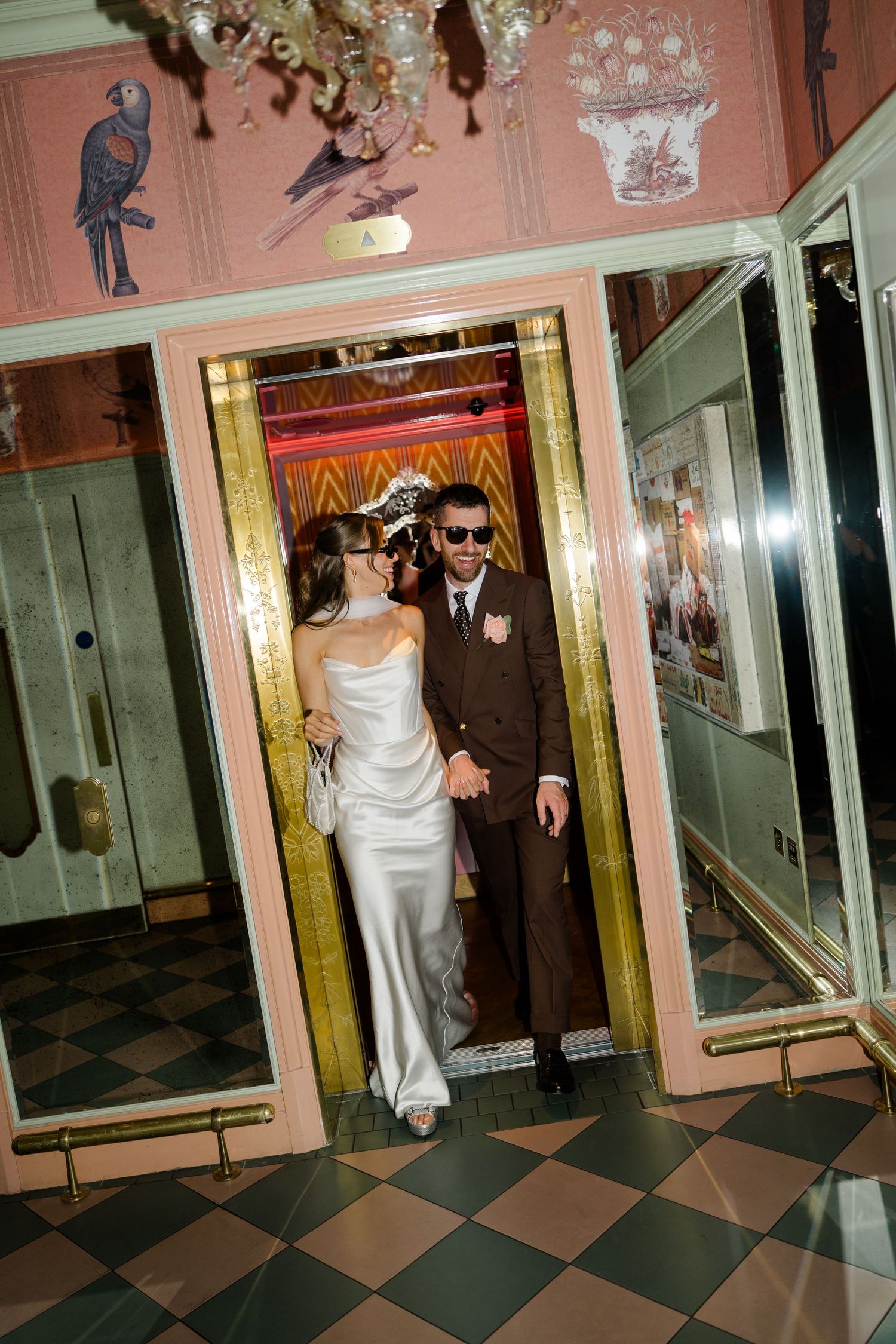 Elegant couple in wedding attire stepping out of a gold elevator in a decorated interior.