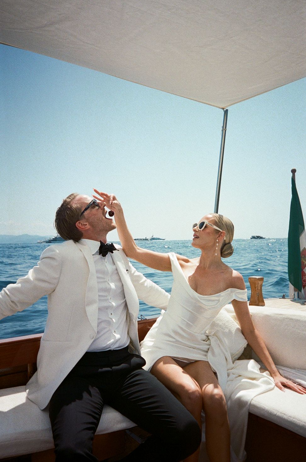 Elegant couple in formal white attire laughing on a luxury wooden boat on the Italian Riviera.