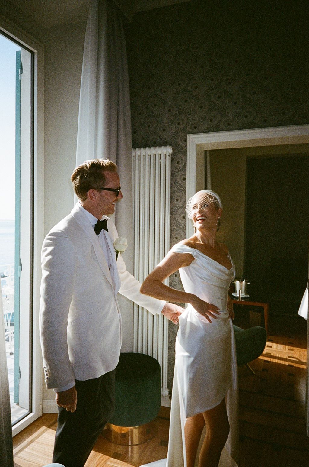 Bride and groom in white wedding attire share a joyful moment in an elegant hotel room with ocean views.