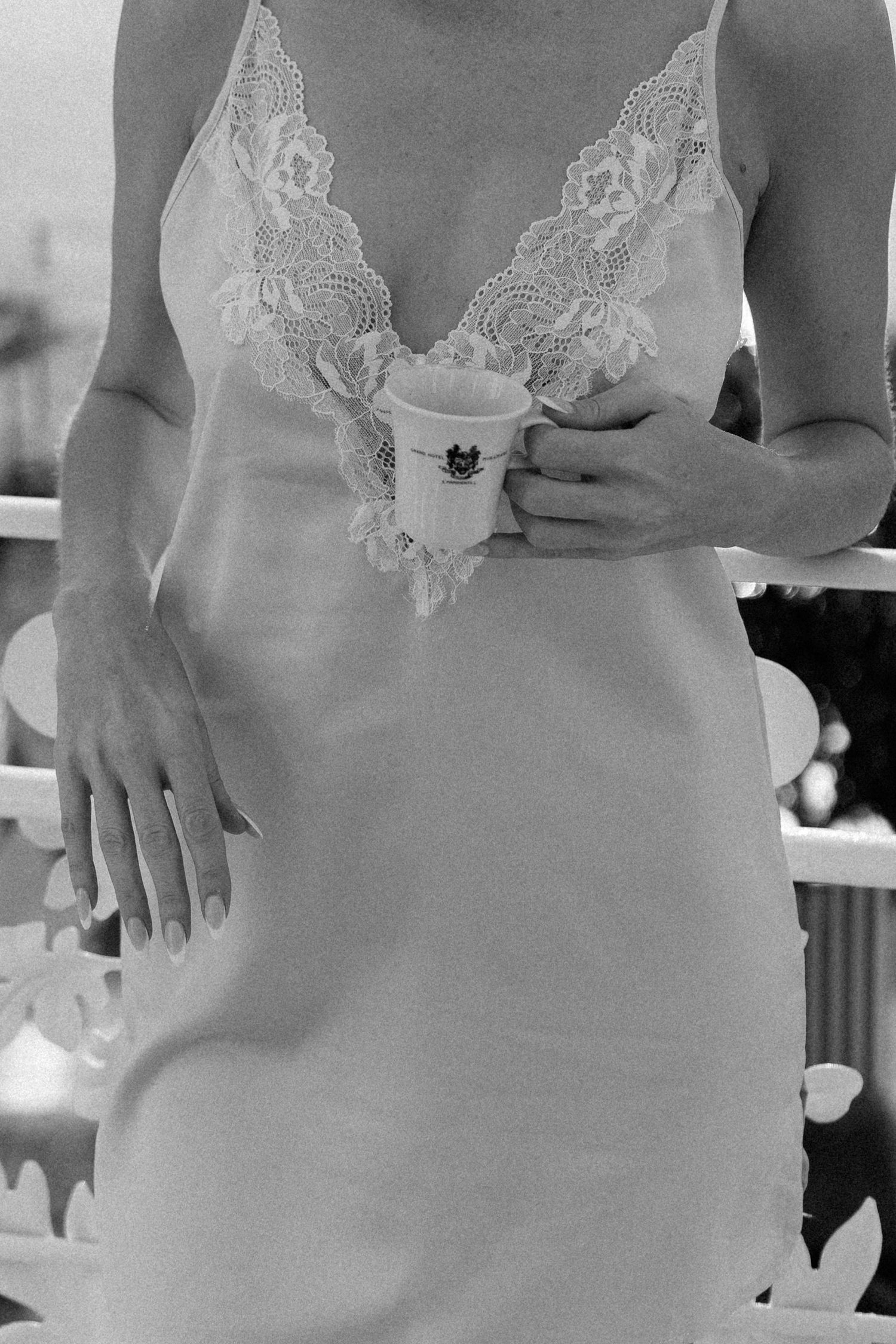 Woman in elegant lace slip dress holds decorative cup on balcony in black and white photo.