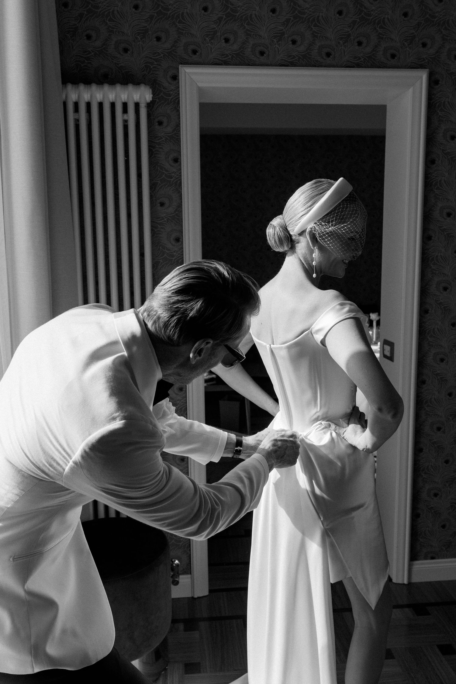 Groom buttoning the back of bride's elegant white wedding dress in a classic interior.