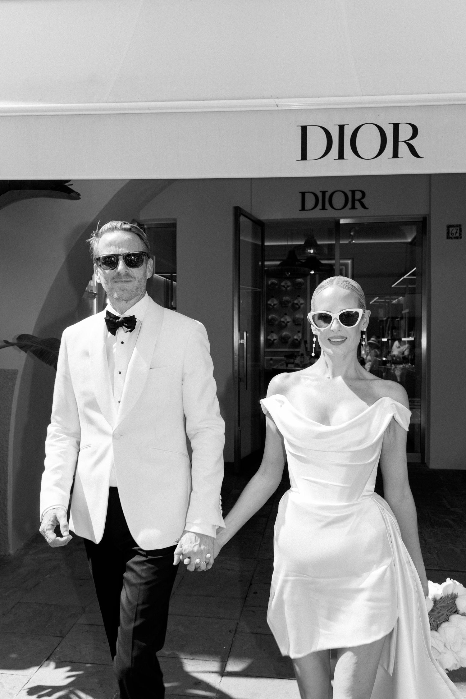 Elegant bride and groom in white wedding attire holding hands outside a Dior boutique in black and white.