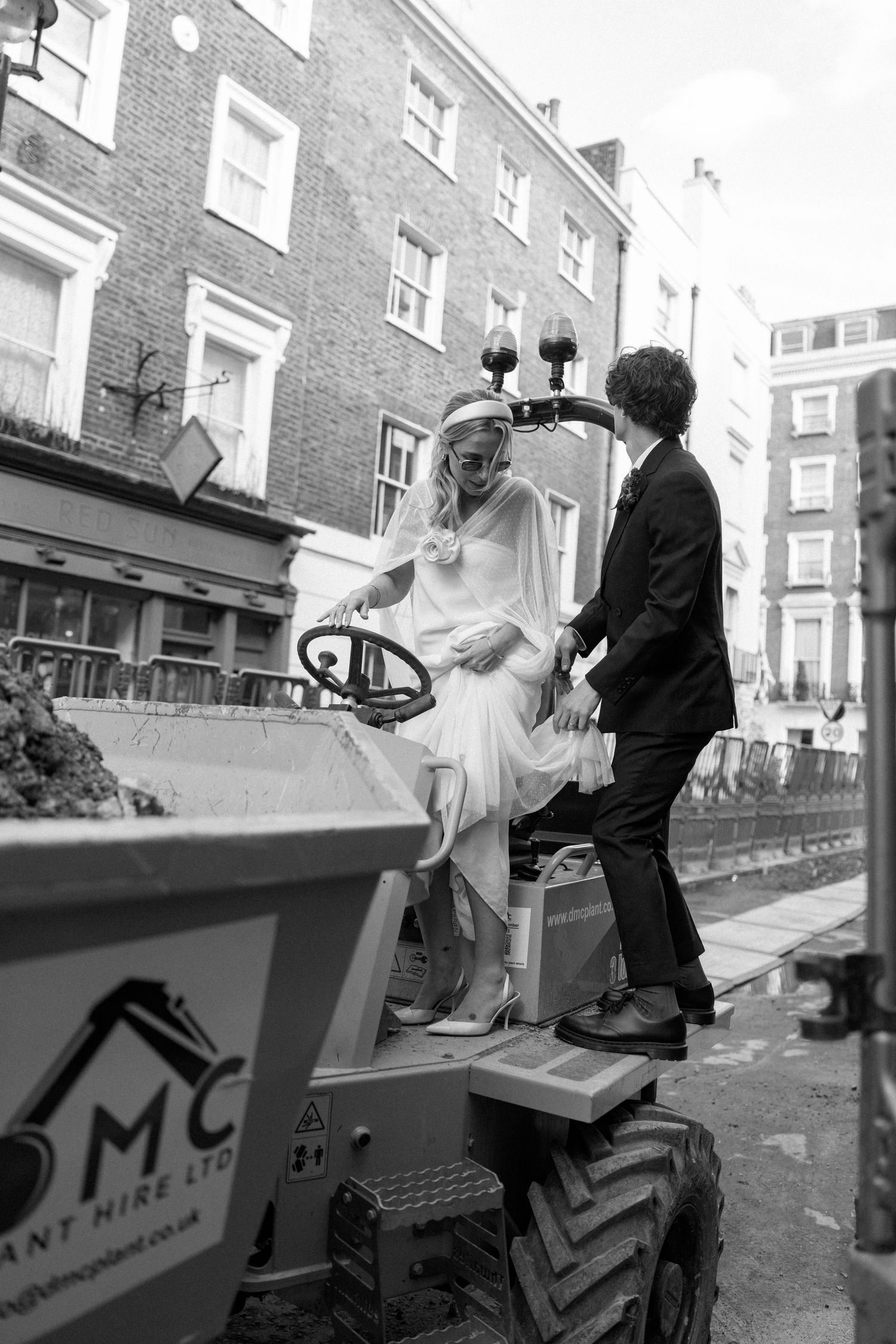 Couple in wedding attire posing playfully near industrial equipment on city street.