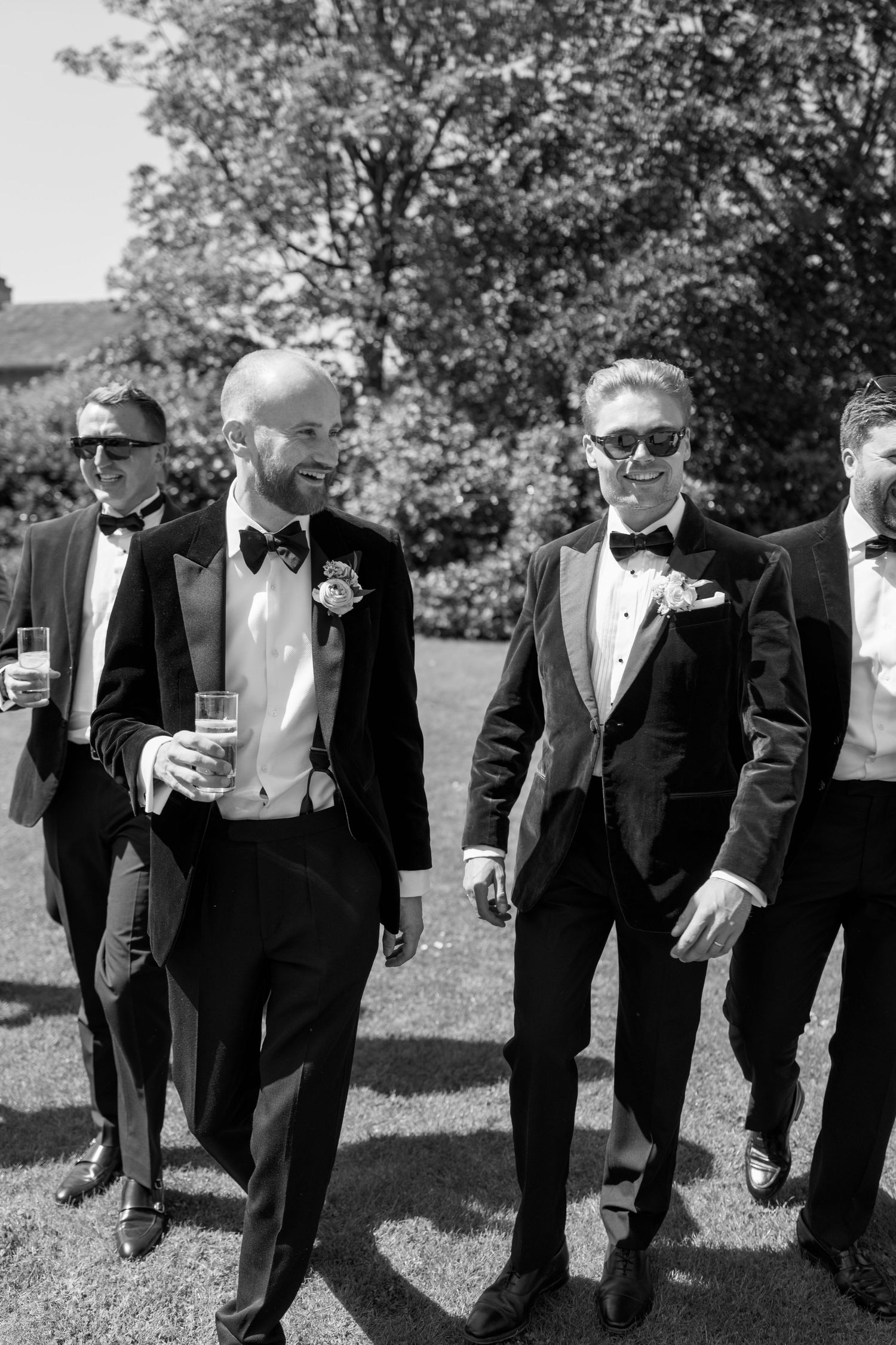 Groomsmen in black tuxedos and bow ties stand in a line on grass while holding drinks.