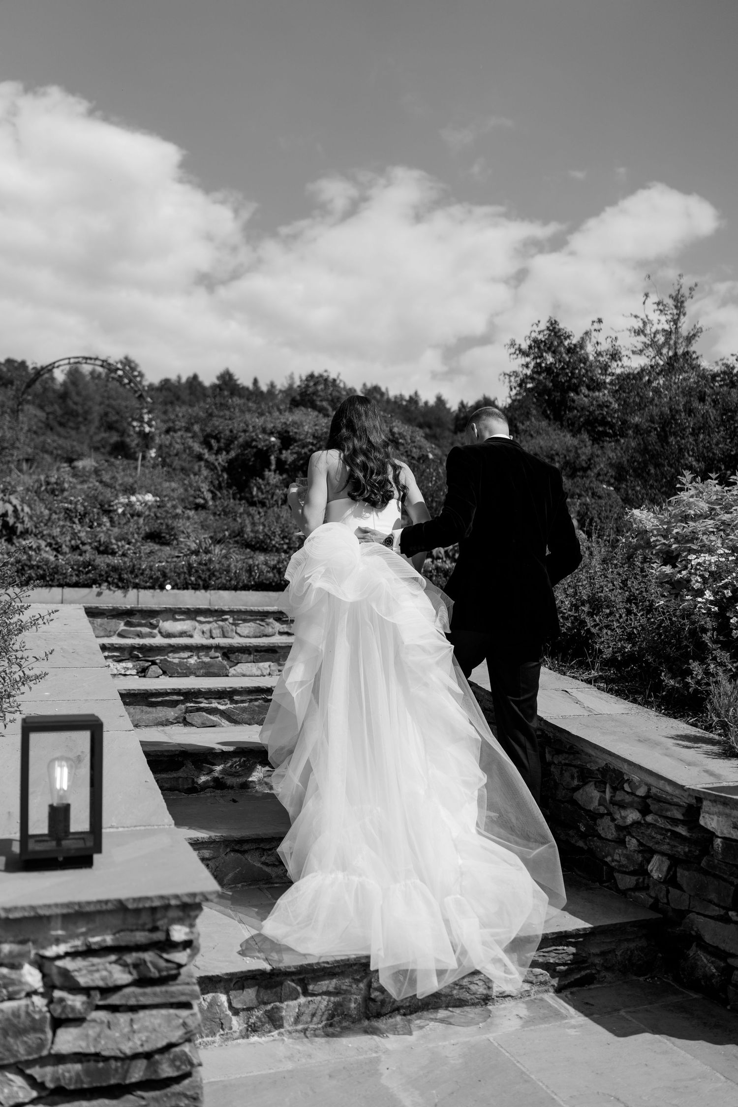 A couple walks along a stone path with a flowing white wedding dress trailing behind.
