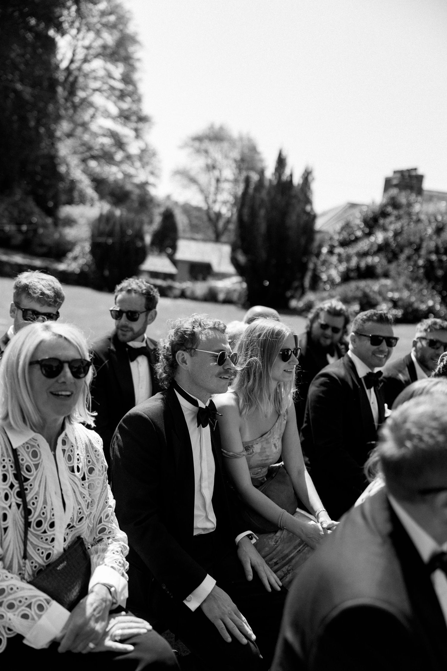 Wedding attendees in sunglasses and formal attire enjoy a sunny outdoor celebration.