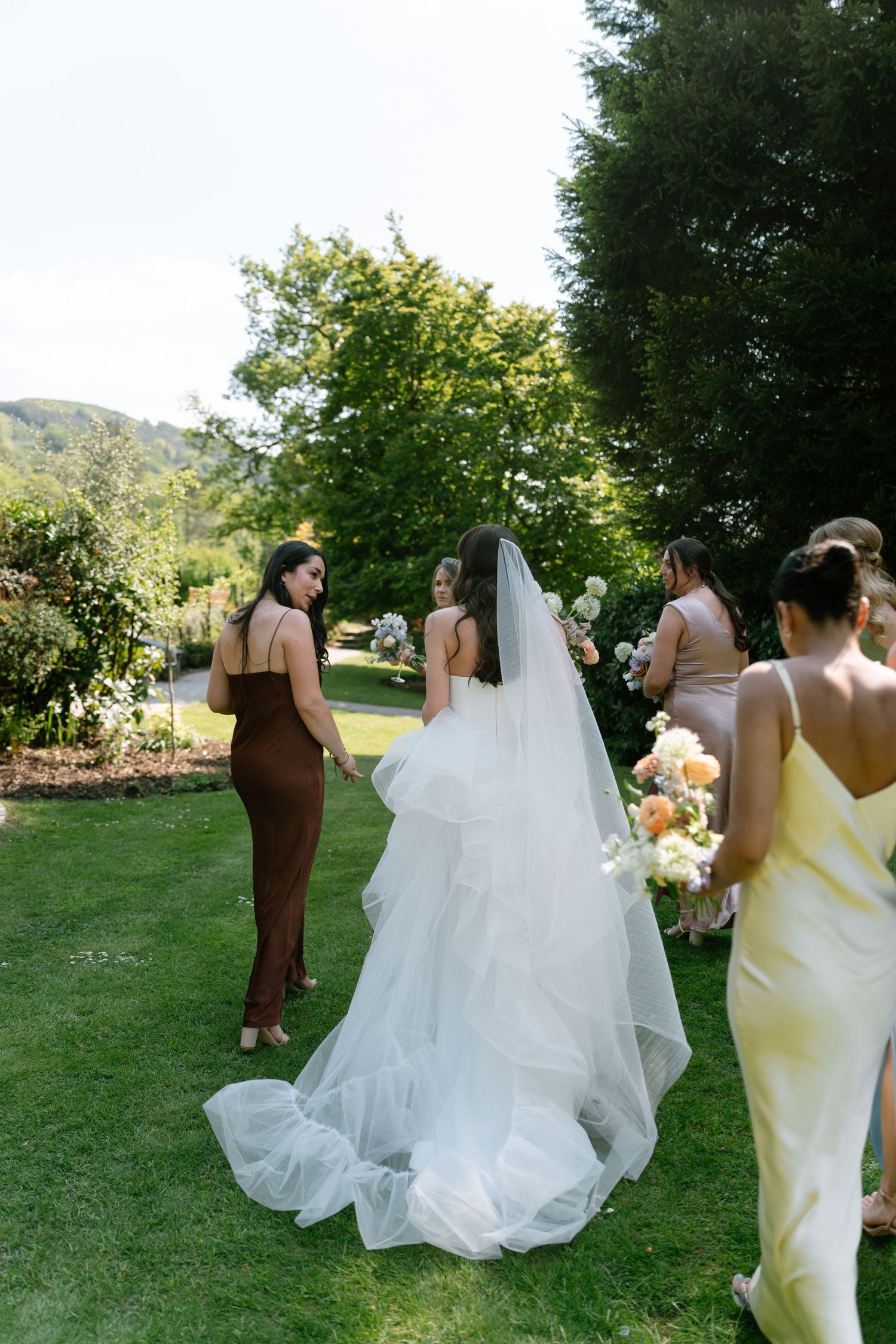 A bridal party in brown and pastel dresses walks across a sunny lawn.
