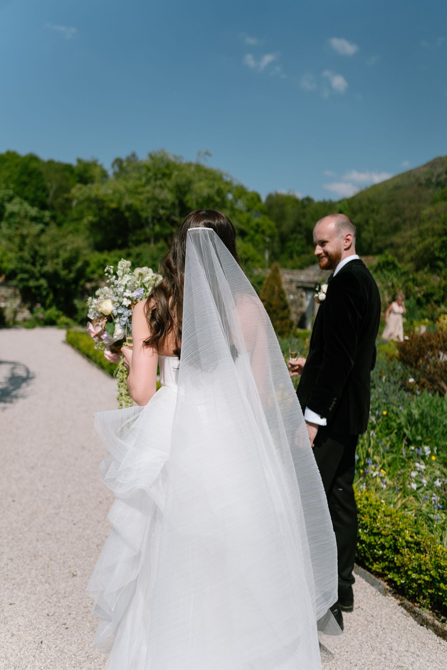 Wedding party walking along a gravel path in a sunny garden setting.