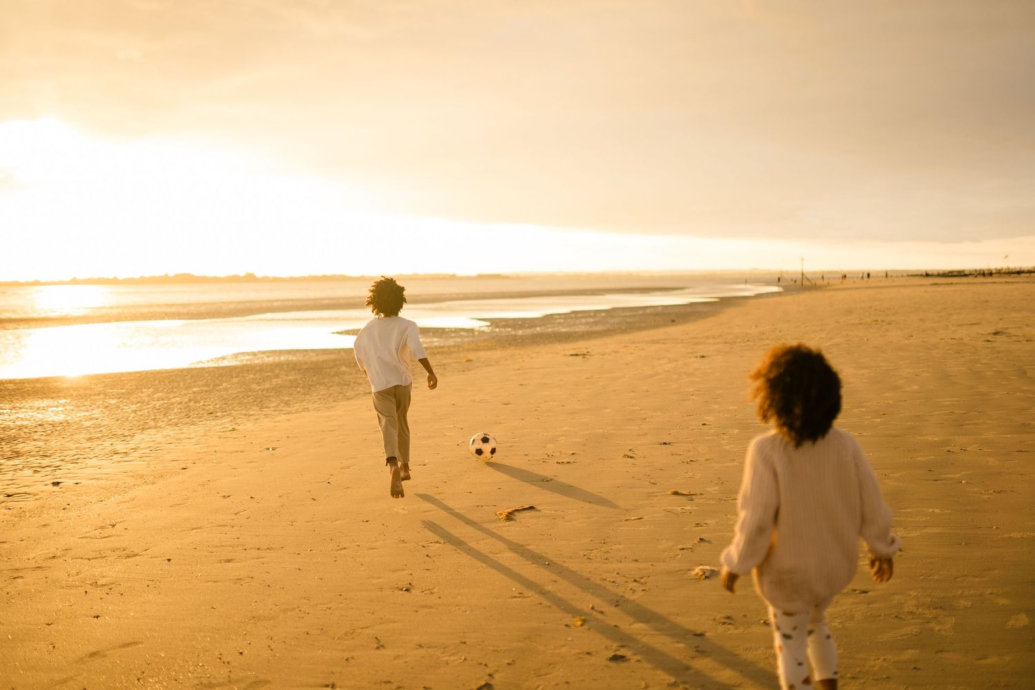 Two figures run along a golden beach at sunset casting long shadows in the sand.