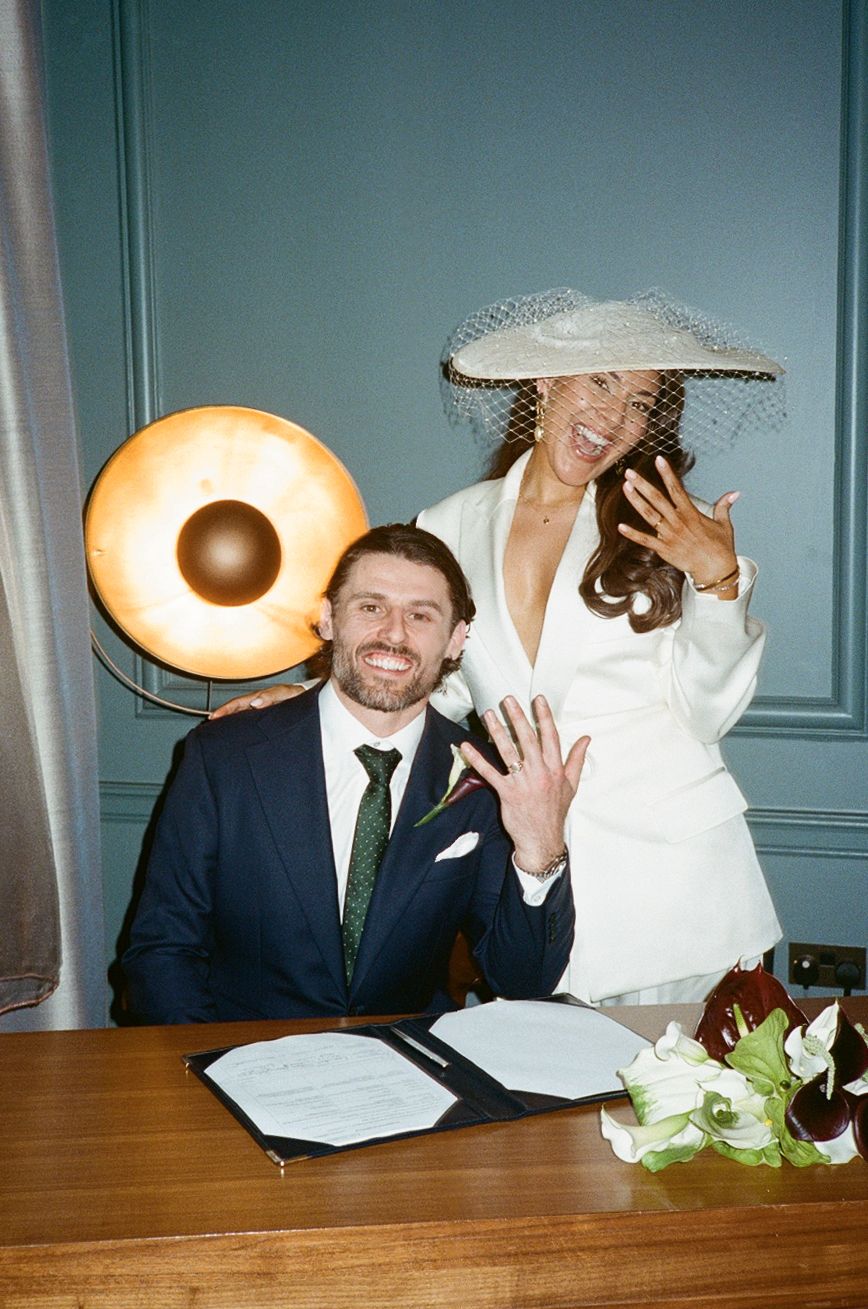 Joyful newlyweds signing marriage certificate, bride in white suit and veil hat showing ring, groom in navy suit smiling.