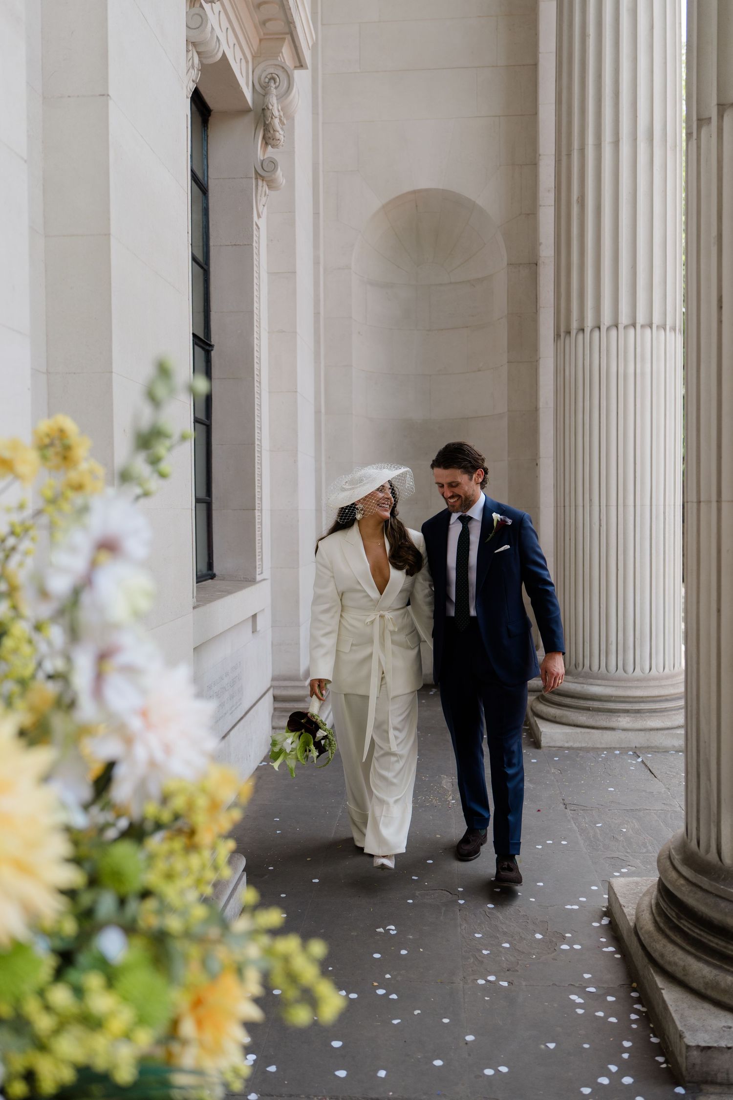 A bride in a white pantsuit and hat walks with a groom in a navy suit between grand stone columns.