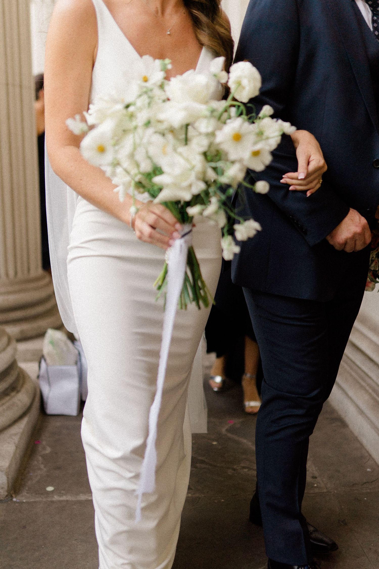 Wedding couple walking with white floral bouquet against formal building backdrop.