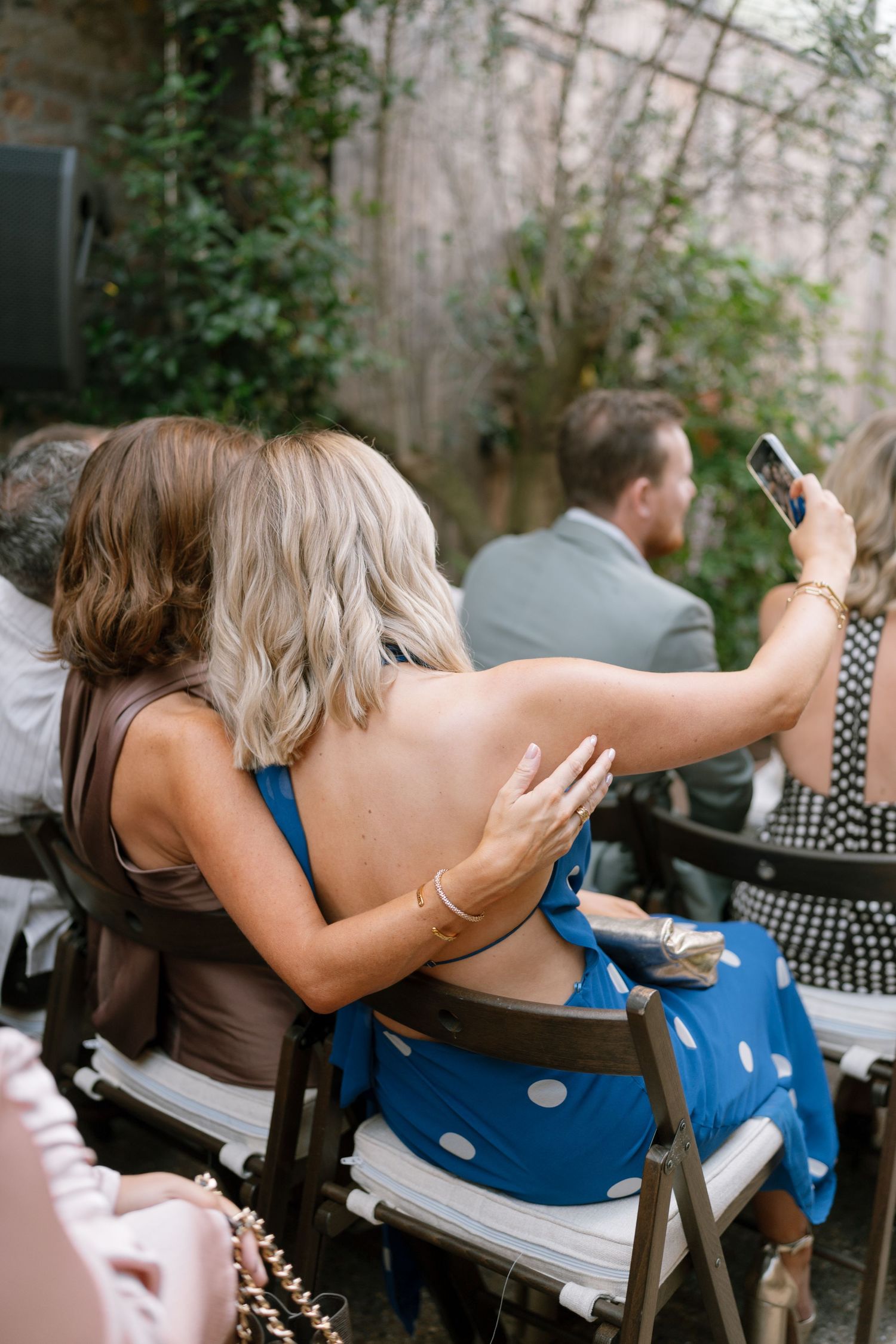 Wedding guests in summer attire seated outdoors looking at their phones during ceremony.
