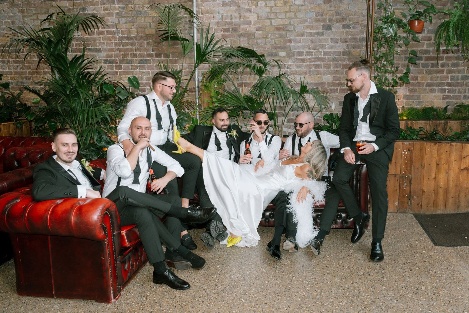 Groomsmen in formal attire relaxing on red leather couch at wedding venue with brick walls.