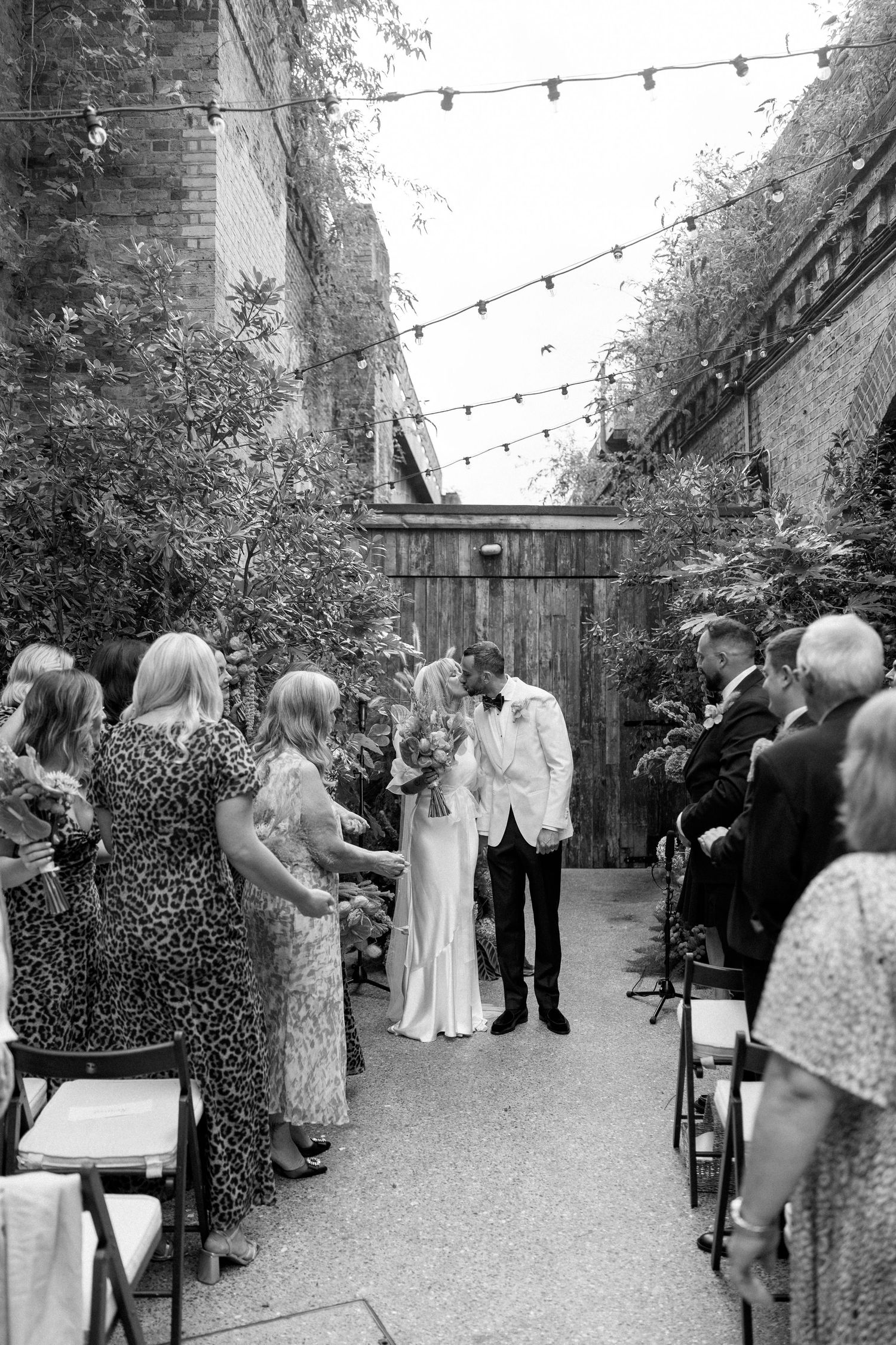Wedding guests gather in an outdoor venue decorated with string lights and greenery.