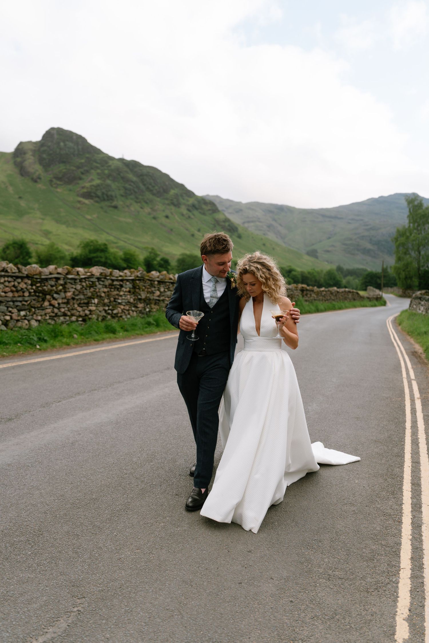 Newlyweds walking down a scenic mountain road with stone walls and green hills.