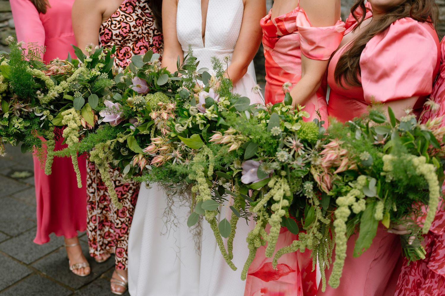 Bridesmaids in coral dresses holding lush greenery bouquets standing in a line.