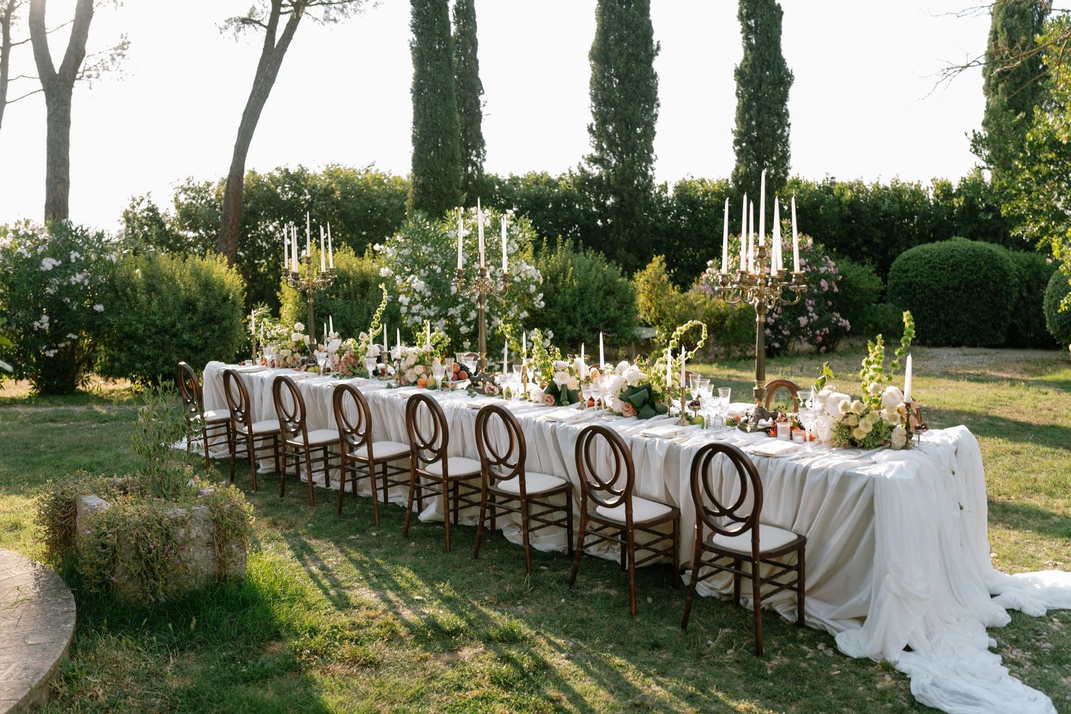 A long outdoor wedding reception table set with white linens and tall candelabras under cypress trees.