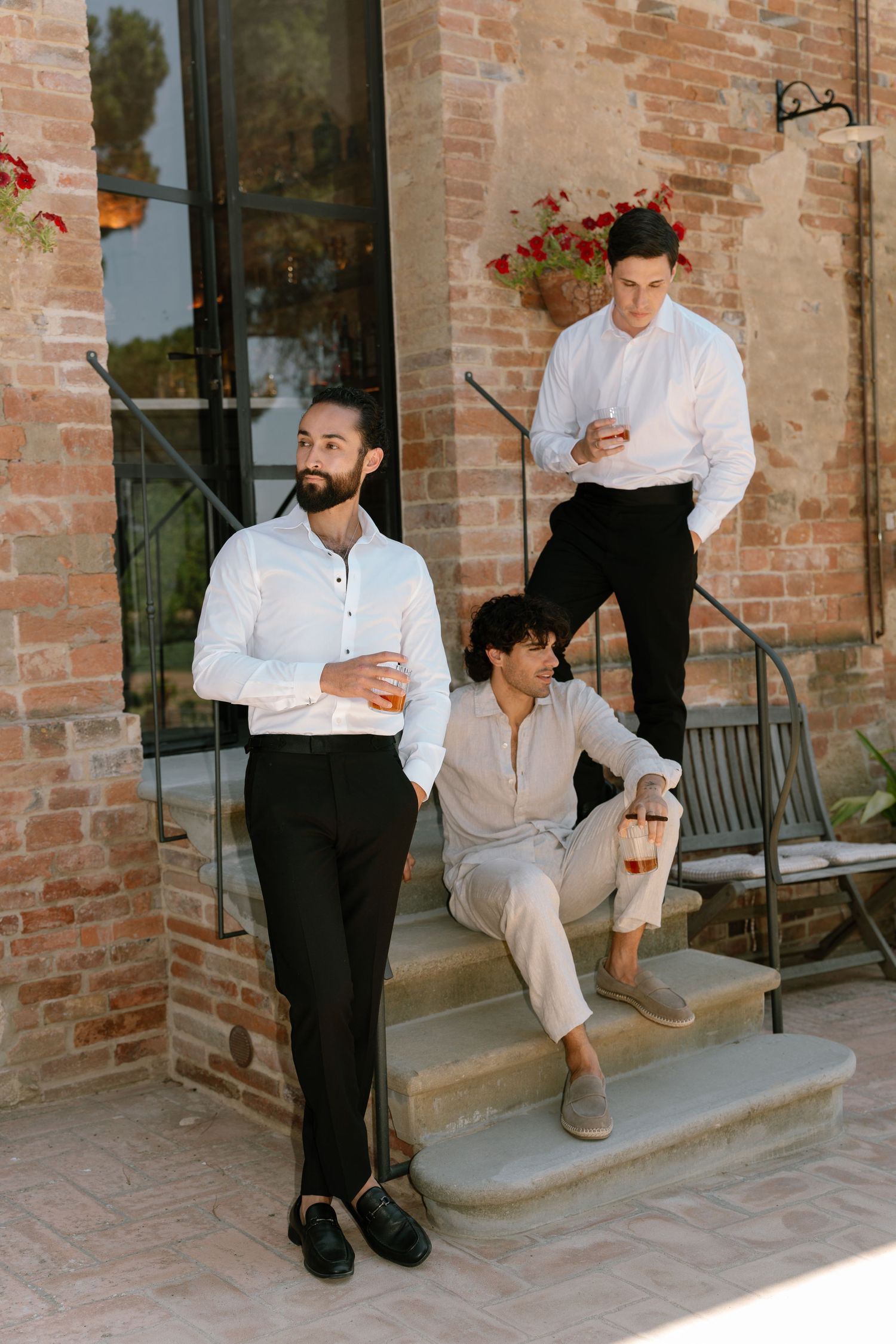 Three people in stylish casual attire pose on brick steps of a rustic building with red flowers in the background.