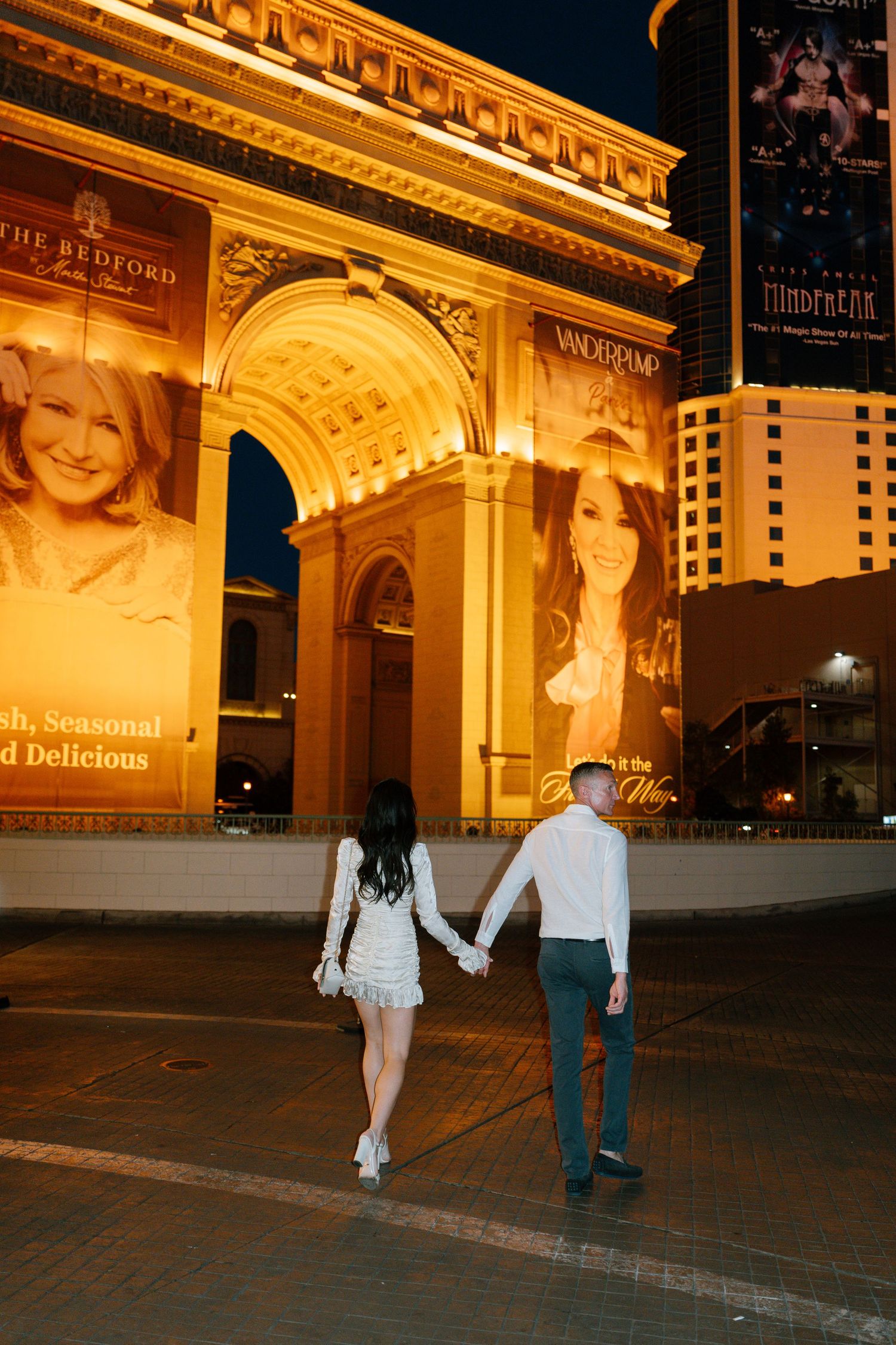 A couple holds hands while walking near an illuminated archway at night in Las Vegas.