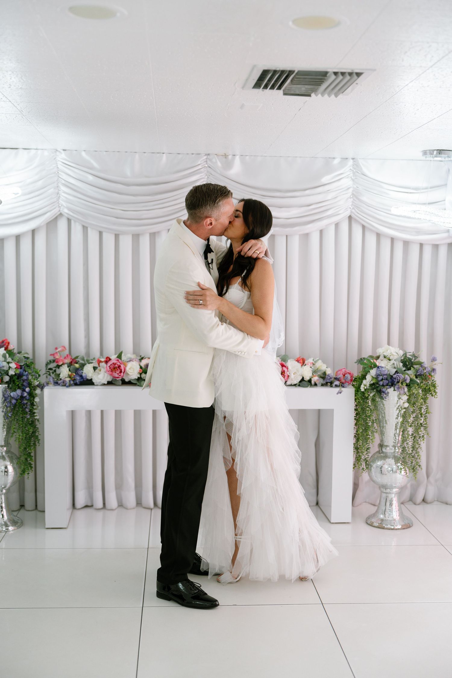 Romantic moment between newlyweds in front of a white floral wedding backdrop.