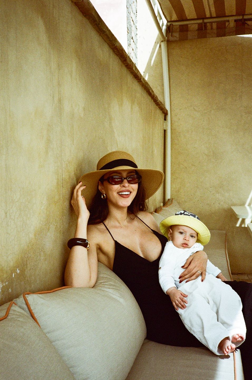 Woman in straw hat and sunglasses smiling while holding a baby wearing a yellow hat on a cozy balcony.
