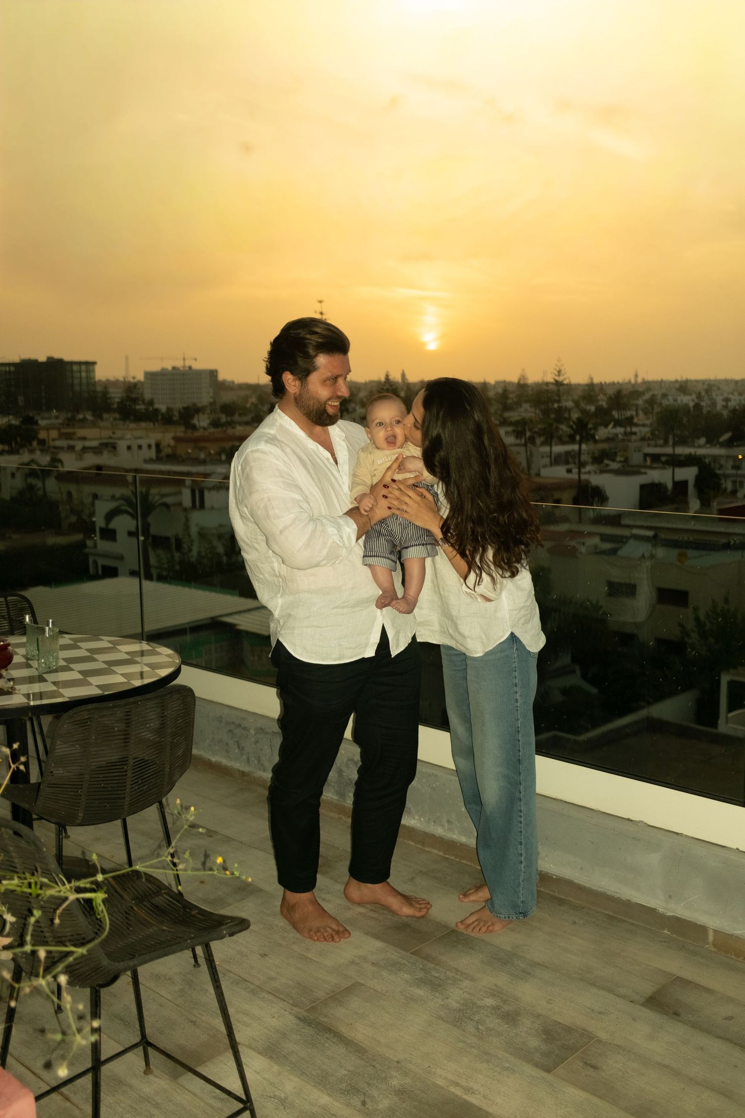 A couple holding their baby on a rooftop terrace at sunset with a city skyline in the background.