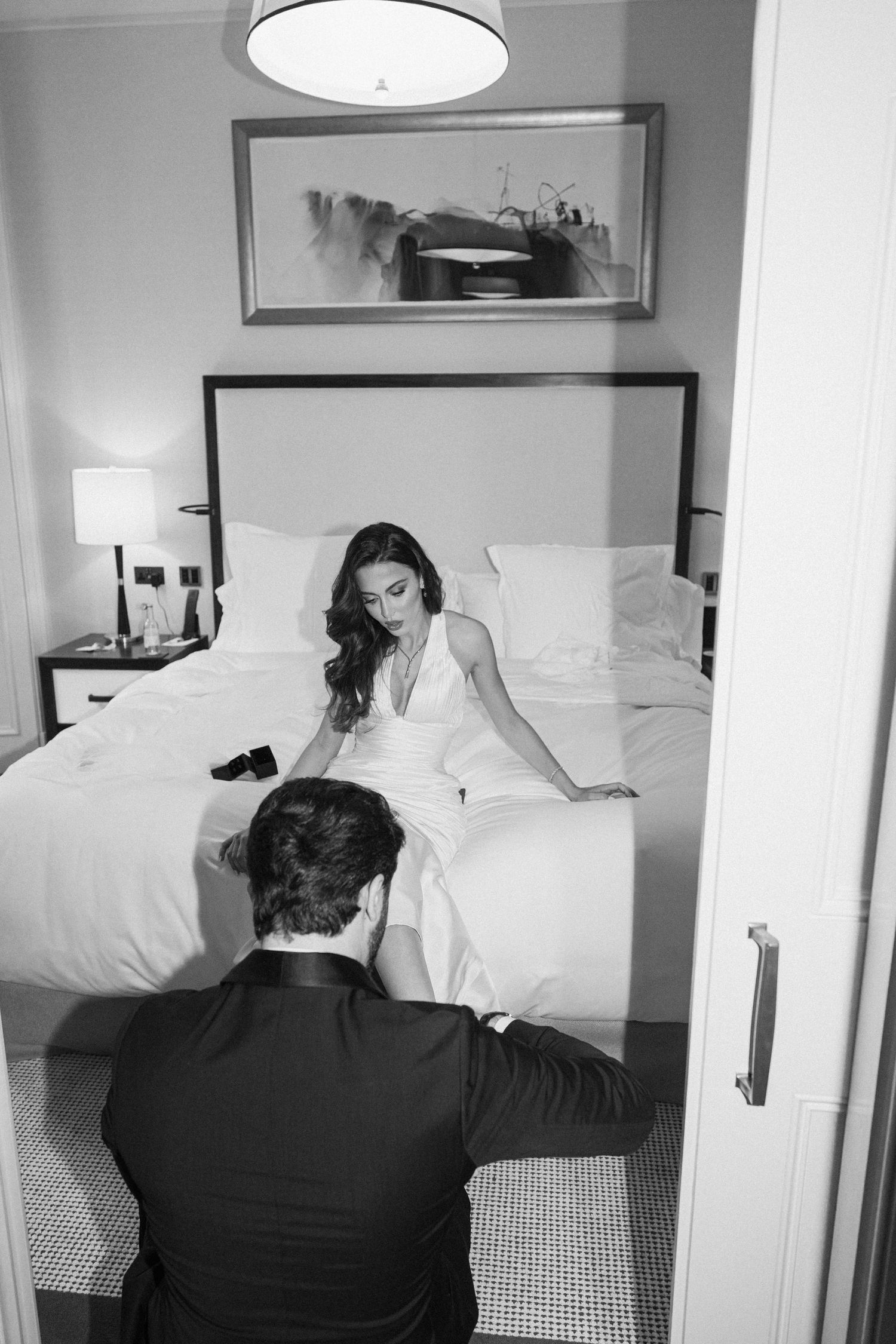 Black and white photo of bride in white gown sitting on hotel bed with groom kneeling before her.